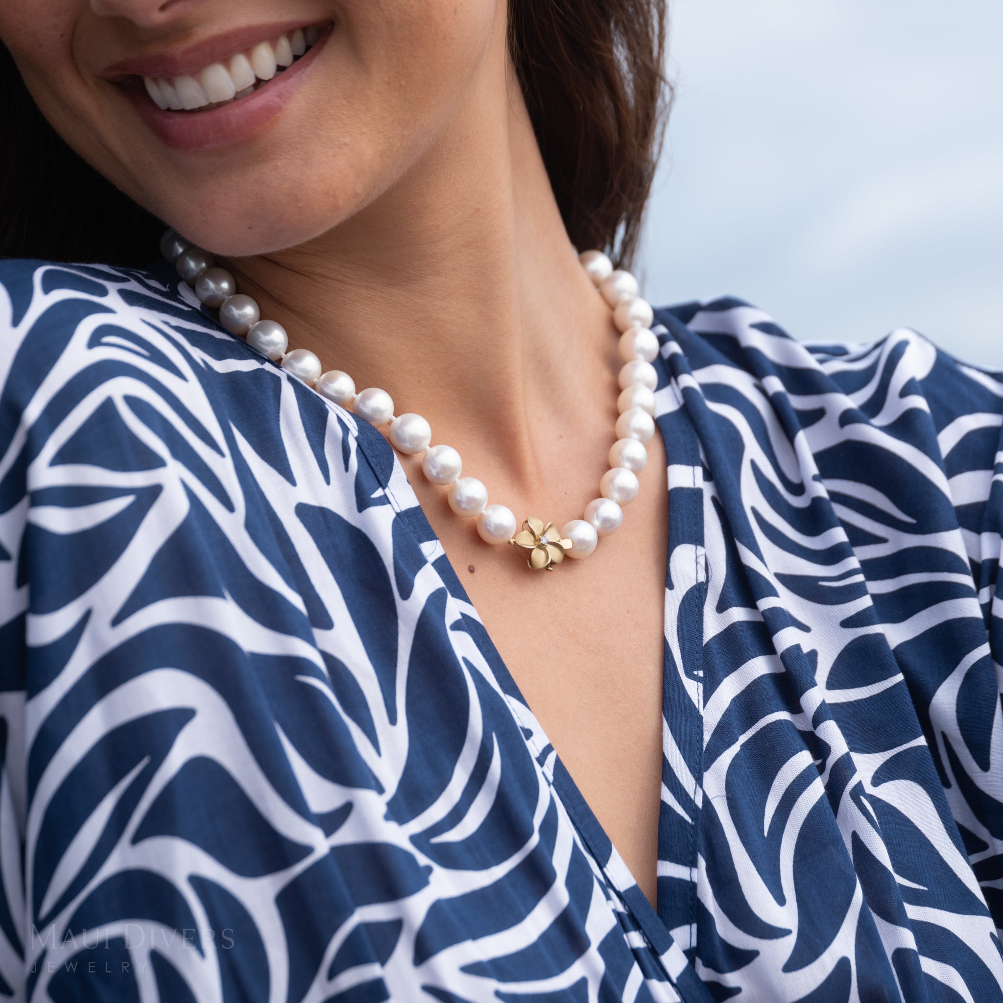 Woman in a blue patterned top wearing a white freshwater pearl necklace with diamond plumeria clasp in yellow gold