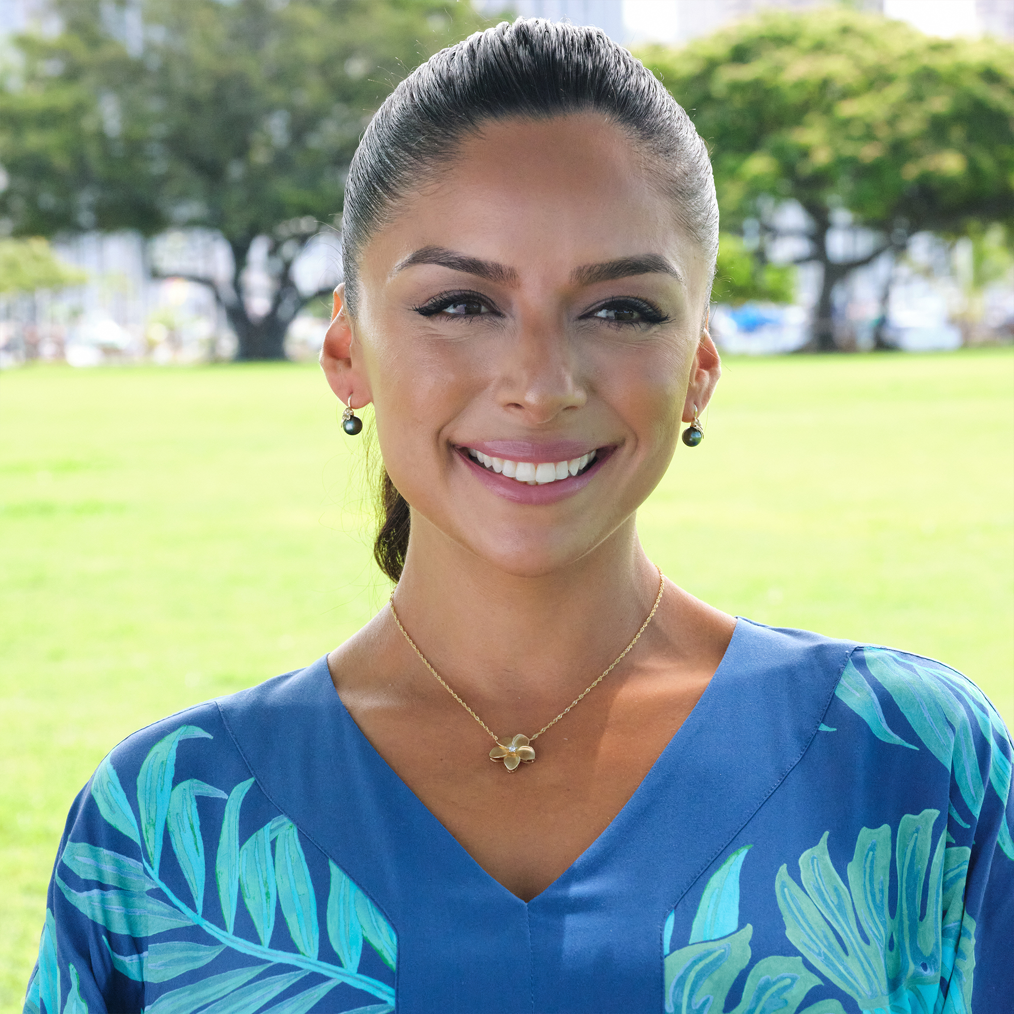 A smiling woman wearing a blue dress wearing a plumeria necklace and plumeria Tahitian black pearl earrings.