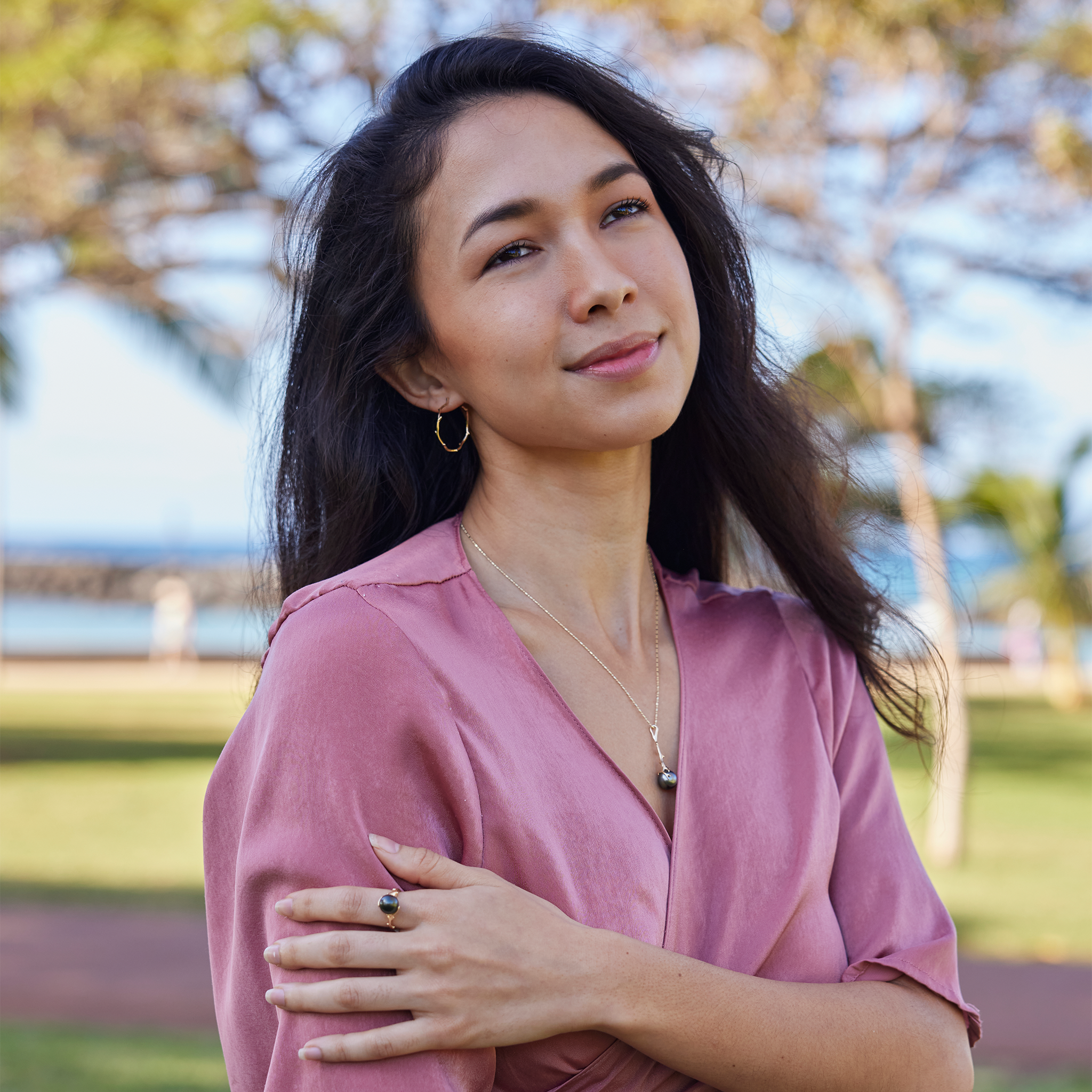 Person in a pink shirt wearing the Heritage Tahitian black pearl pendant in 14k yellow gold on a bar-ball chain and matching hoop earrings