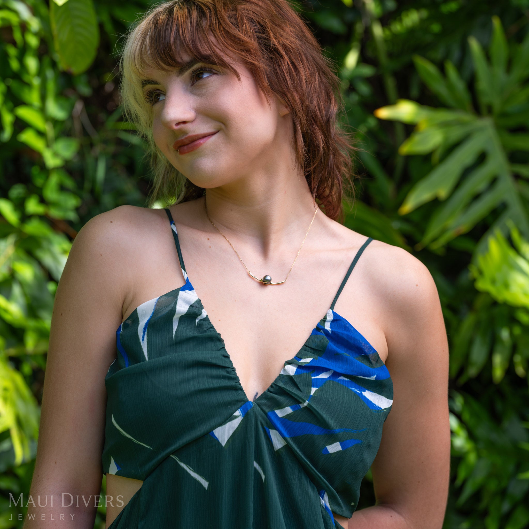 A lifestyle shot of a woman wearing a gold necklace with a curved heritage collection black coral-inspired design with a Tahitian black pearl in the middle, paired with a blue and green patterned top against a soft-focus tropical background
