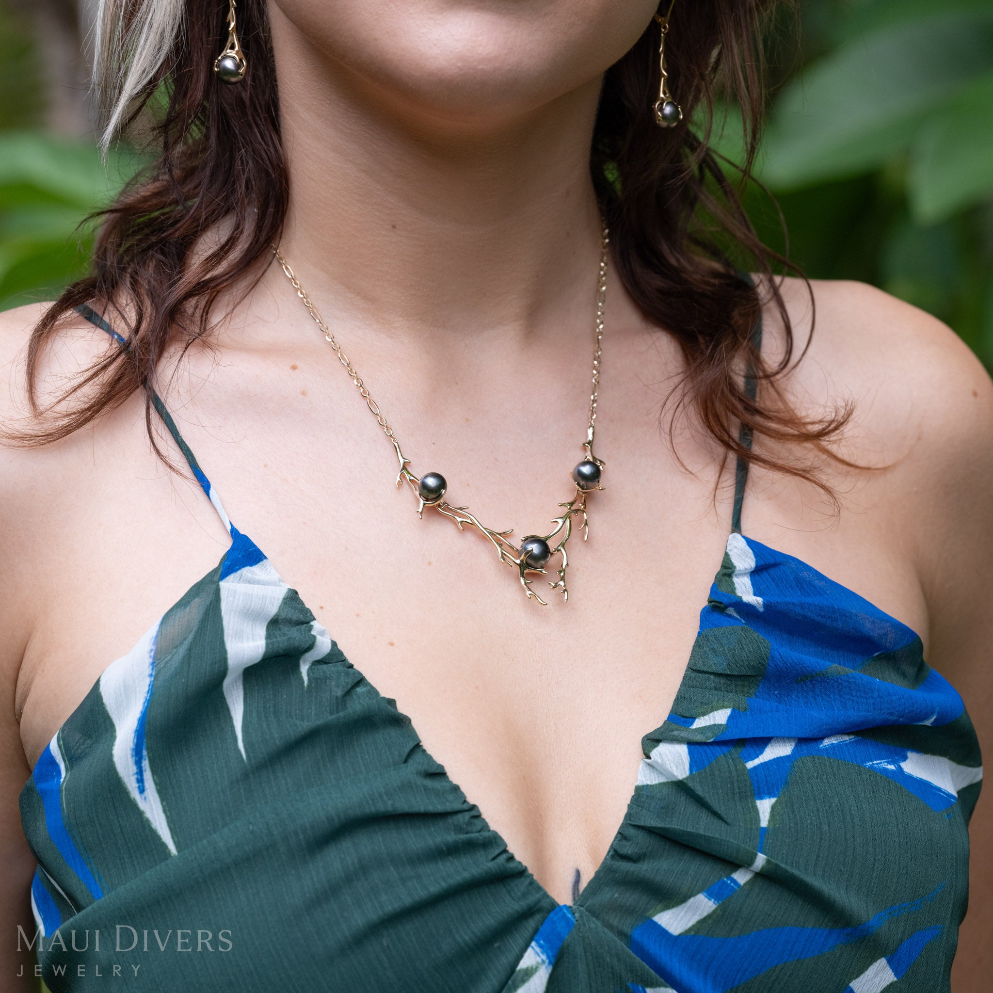 A close-up lifestyle shot of a model wearing a Tahitian black pearl necklace with black coral-branch inspired designs in 14k yellow gold, along with matching dangling earrings. The model is wearing a blue and green patterned top against a soft-focus green background.
