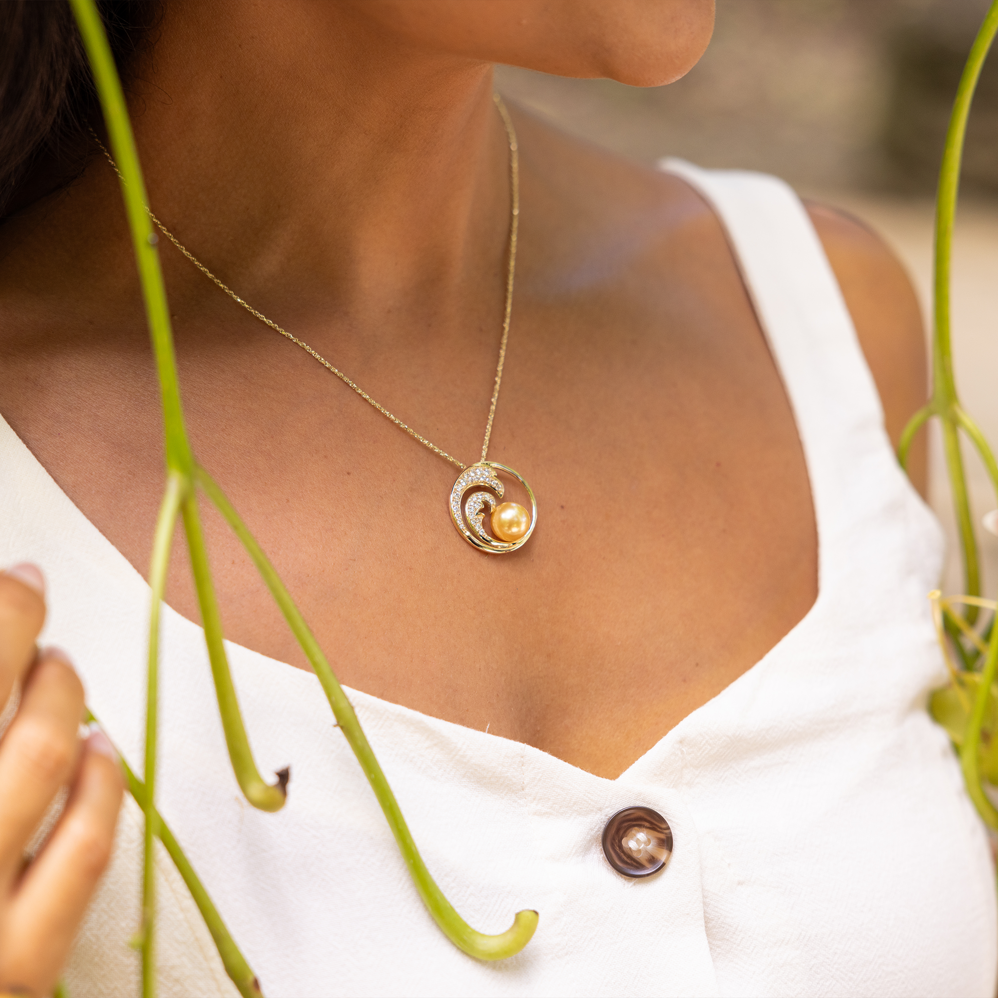 Close up of a woman in a white dress wearing a gold diamond wave South Sea gold pearl pendant on a box chain with green vines framing her