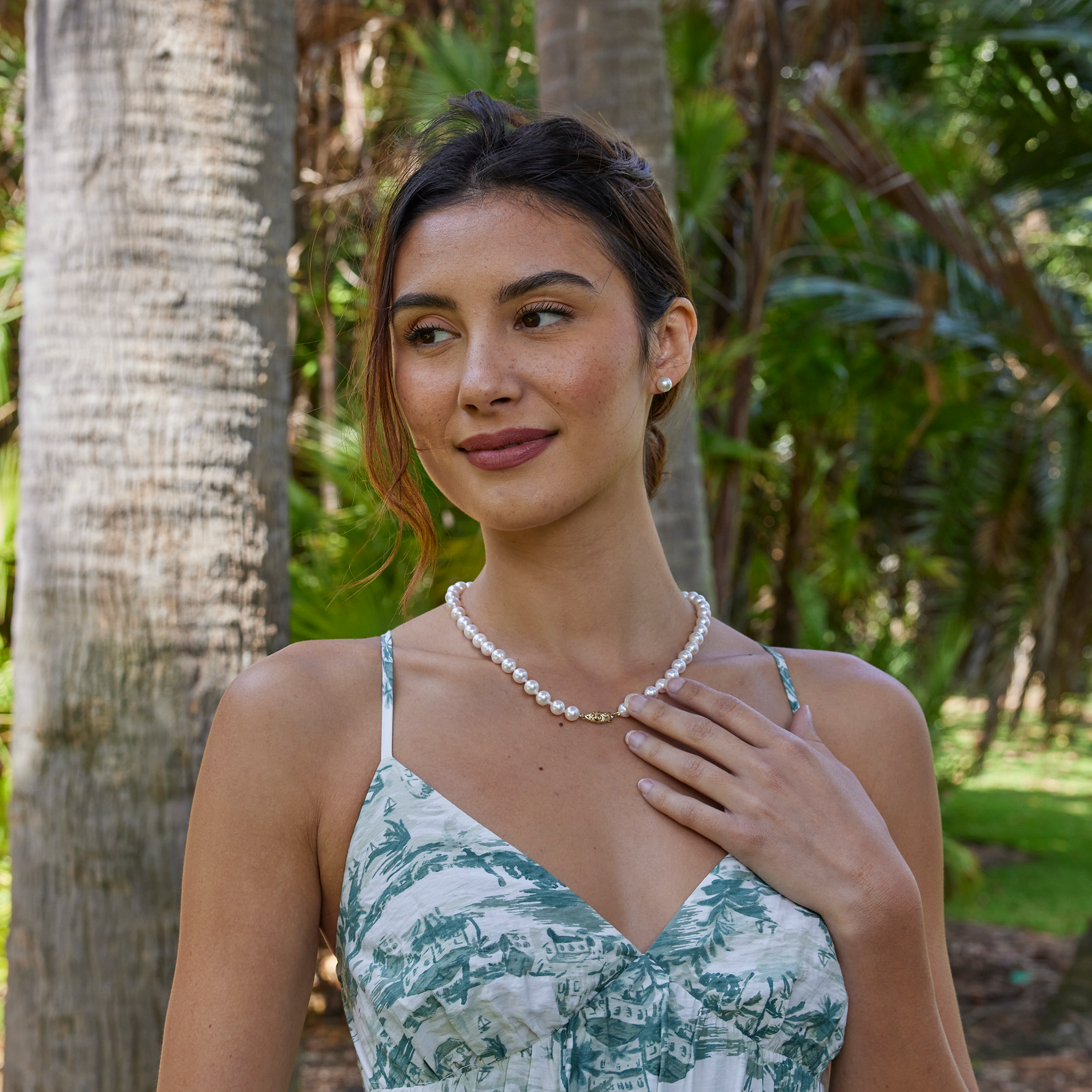 Smiling woman wearing an Akoya white pearl strand necklace with a 14k gold clasp and matching pearl earrings against a blurred background