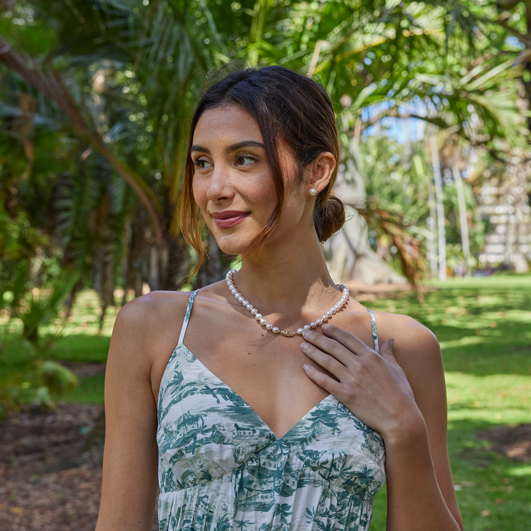 Smiling woman wearing an Akoya white pearl strand necklace with a 14k gold clasp and matching pearl earrings against a blurred background