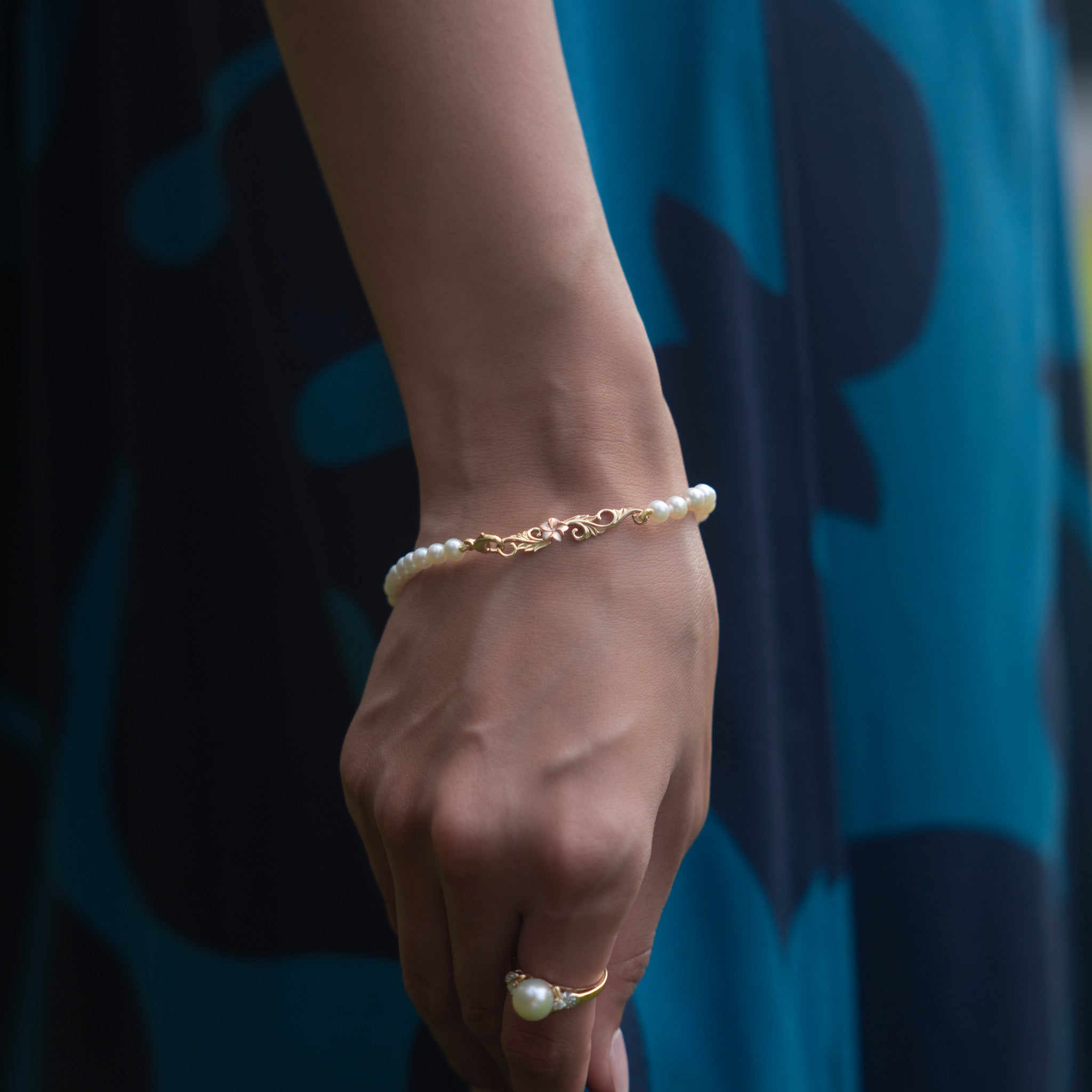 Close-up of a woman wearing a freshwater white pearl bracelet with 14k yellow gold Hawaiian Heirloom scrollwork and a 14k rose gold plumeria clasp
