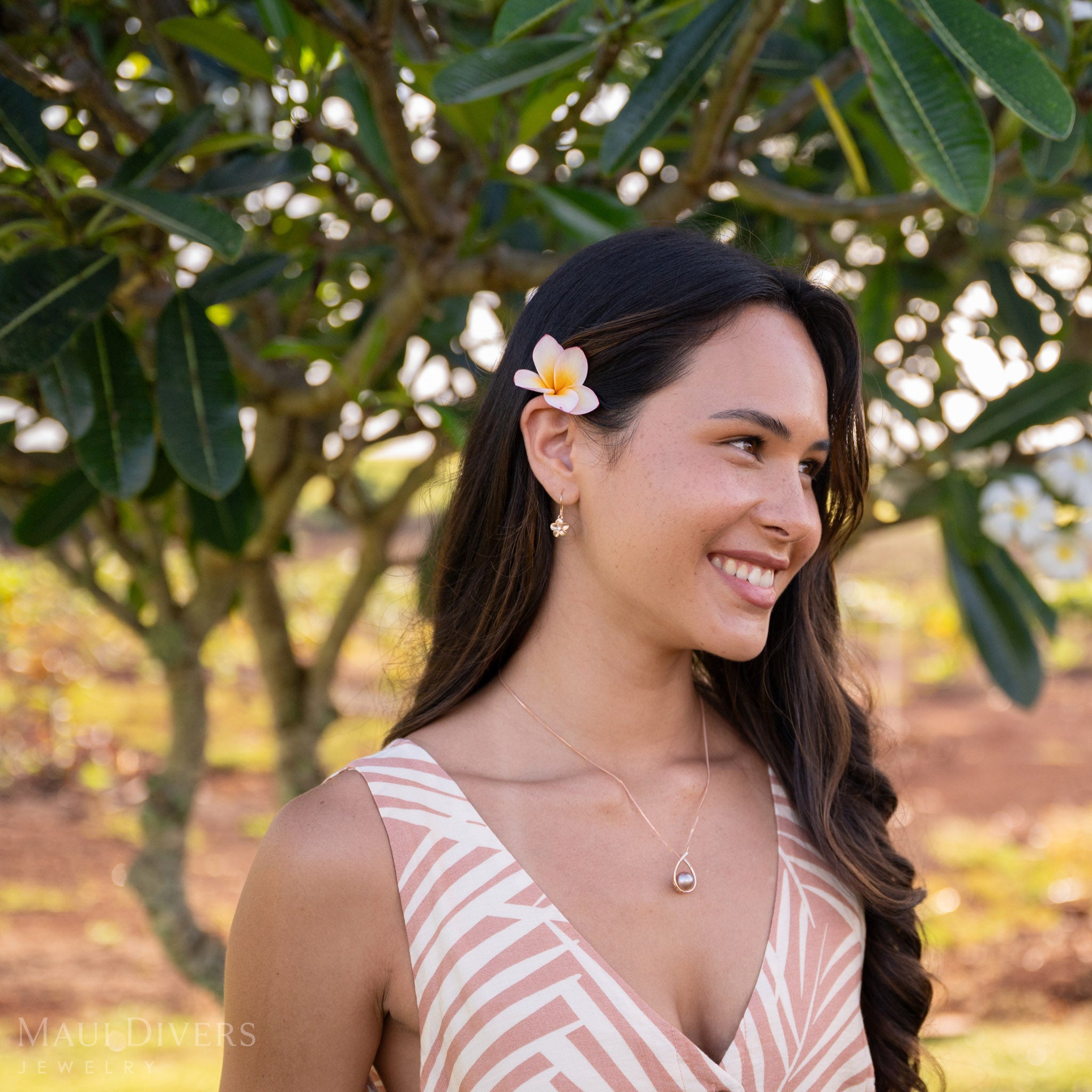 A smiling woman with a plumeria flower in her hair wearing a Plumeria Earring with a diamond in 14k rose gold, in front of a blurred plumeria tree