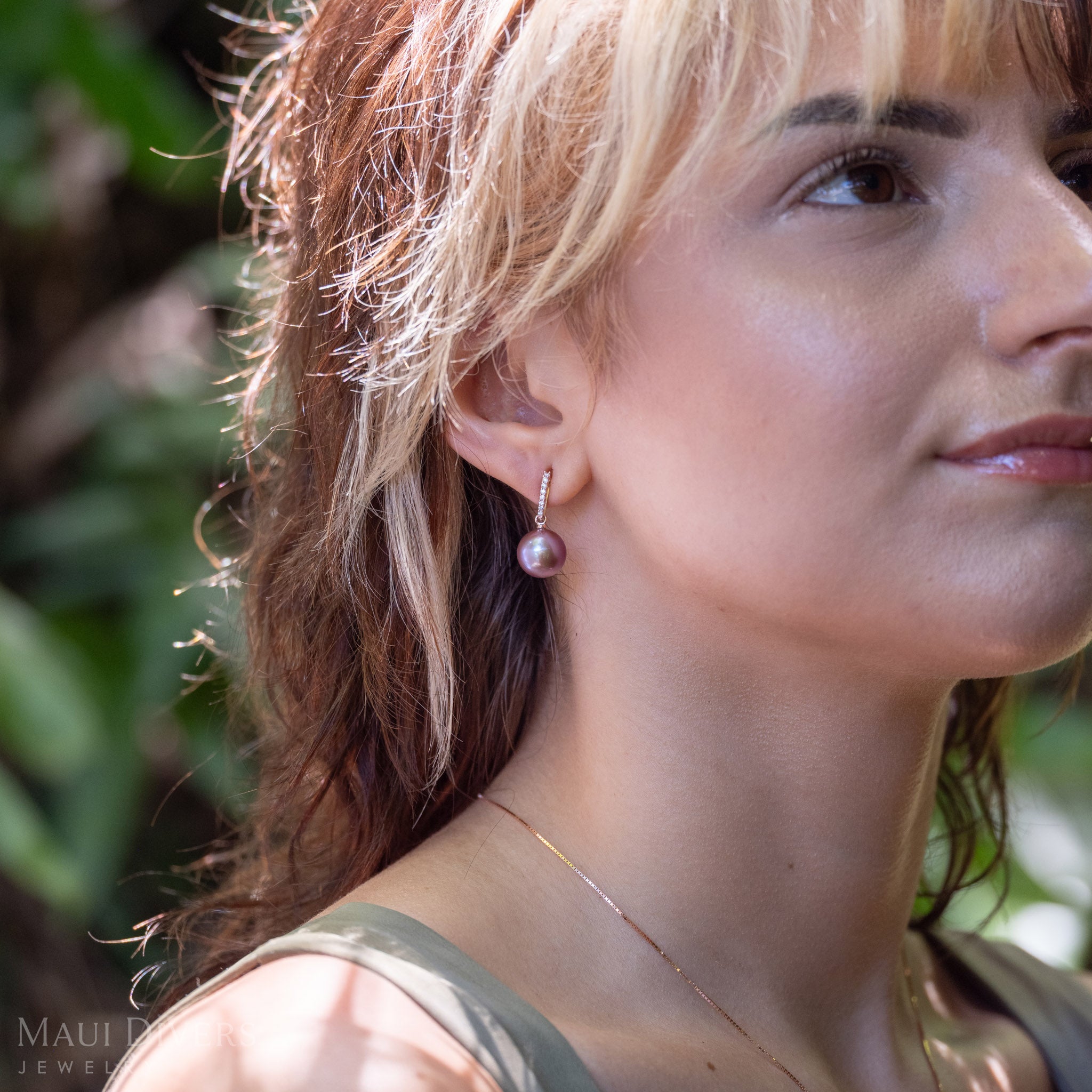 A close-up side profile of a woman wearing a single dangling ultraviolet freshwater pearl diamond drop earring in 14k rose gold, set against a blurred green outdoor background.