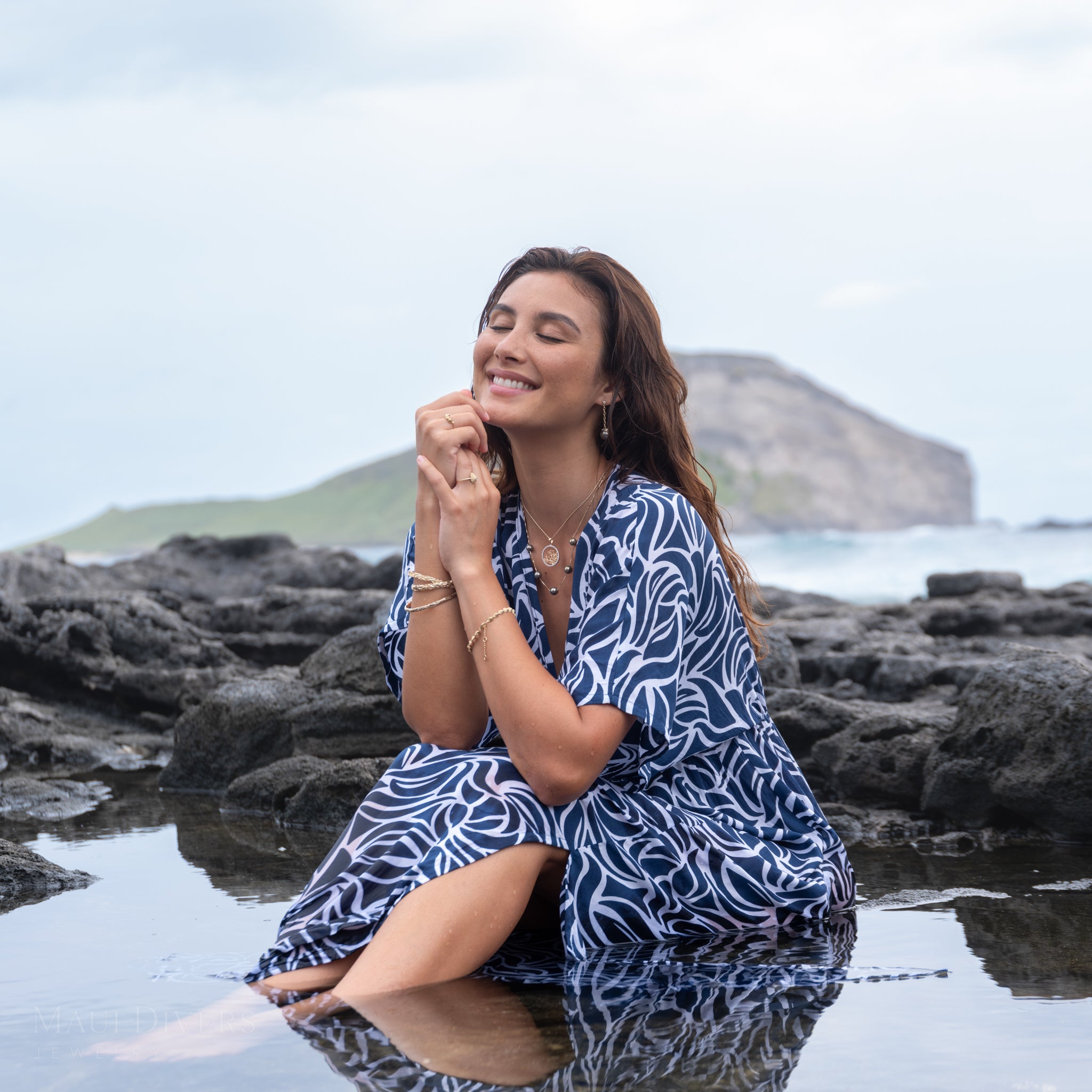Woman wearing the Reefs Starfish Tahitian Black Pearl Earrings in Gold with Diamonds in a blue patterned dress, sitting on rocks by the water