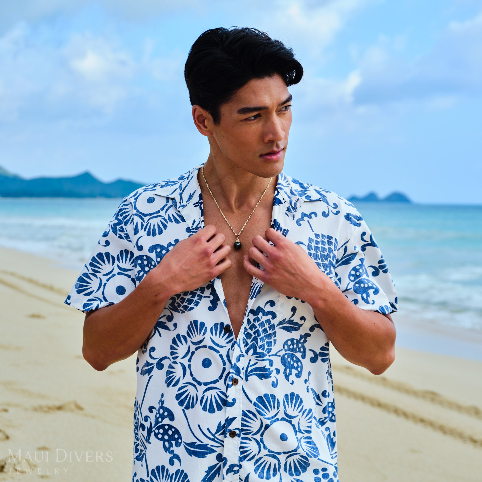 Man wearing a blue and white patterned shirt with a Fish Net Tahitian Black Pearl Pendant in Gold on a beach