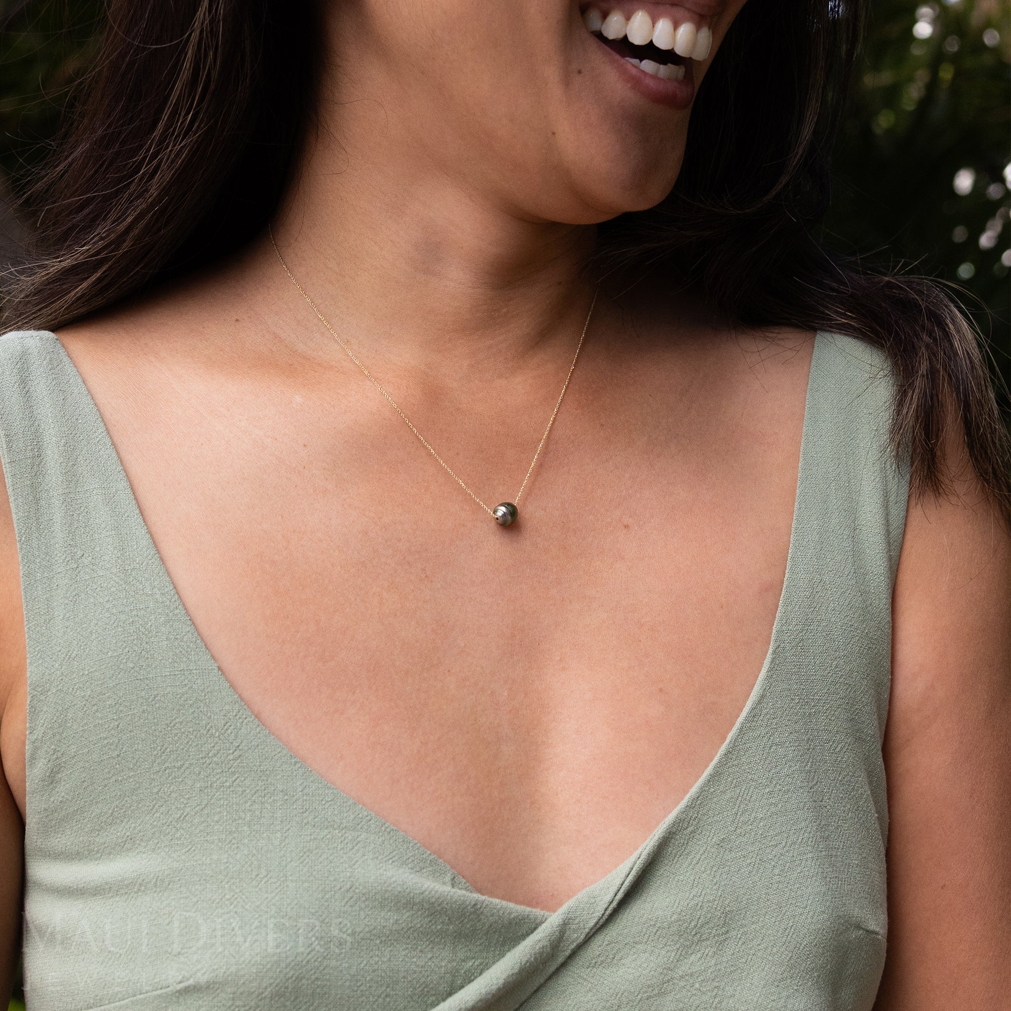 Close-up of a smiling woman wearing an Adjustable Tahitian Black Pearl Necklace in 14k yellow gold against a blurred background