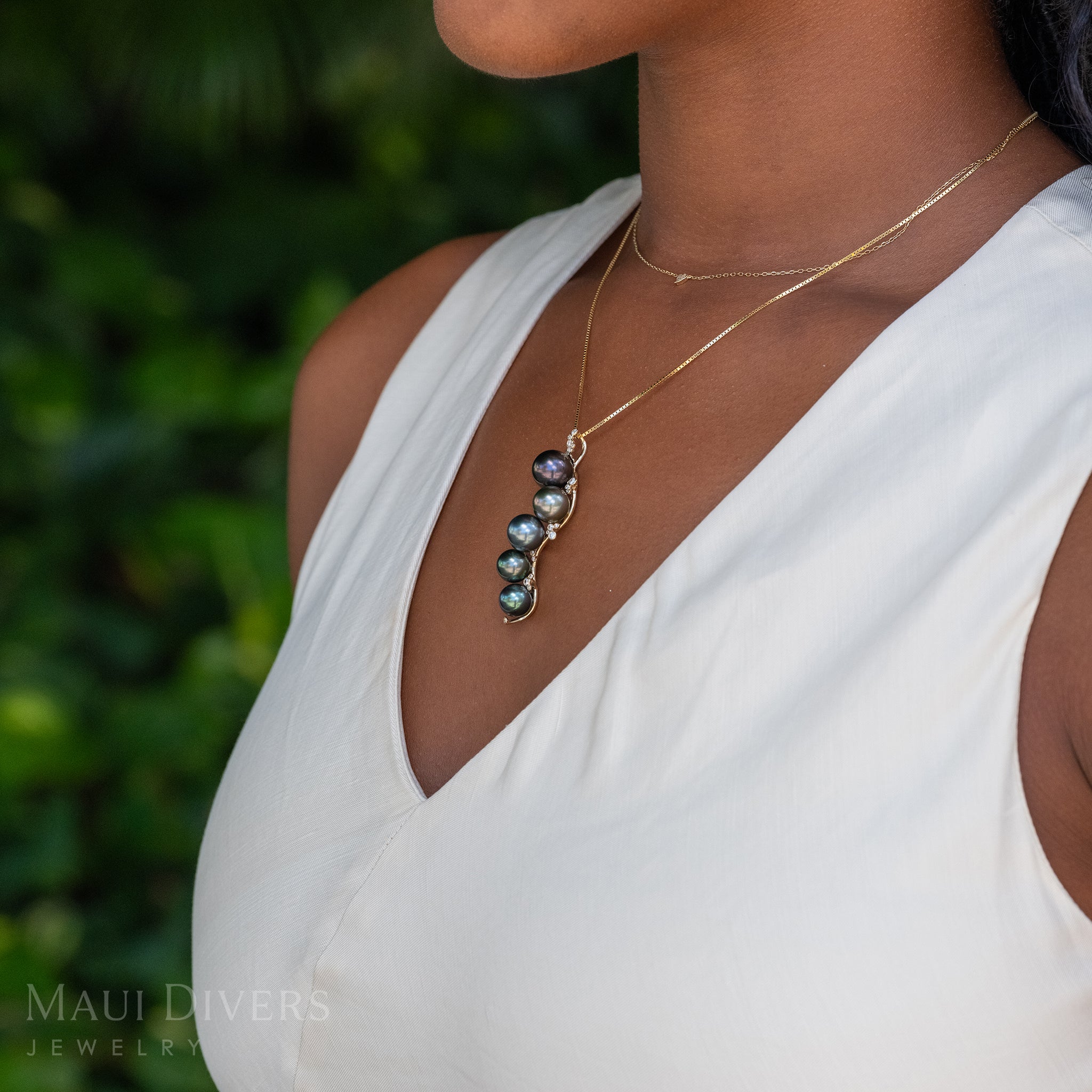 Side-view of a woman in a white top wearing a Bubbling Tahitian Black Pearl Pendant in 14k yellow gold with diamonds on a box chain, layered with a tiny diamond heart necklace, against a blurred leafy green background
