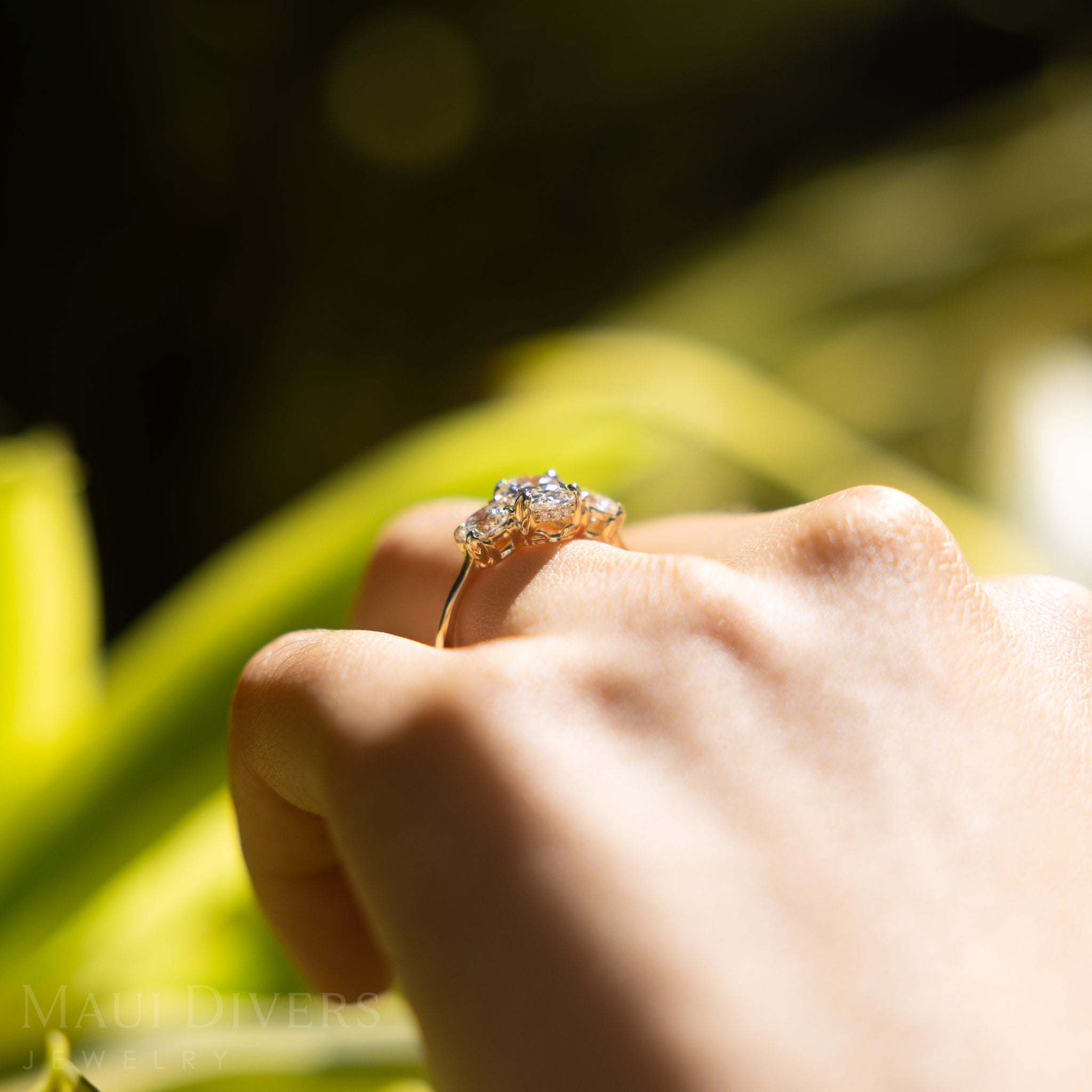 Hand wearing a Hidden Hawaiʻi Maile Three Stone Lab Grown Diamond Ring in 14k Yellow Gold, held against a blurred natural background