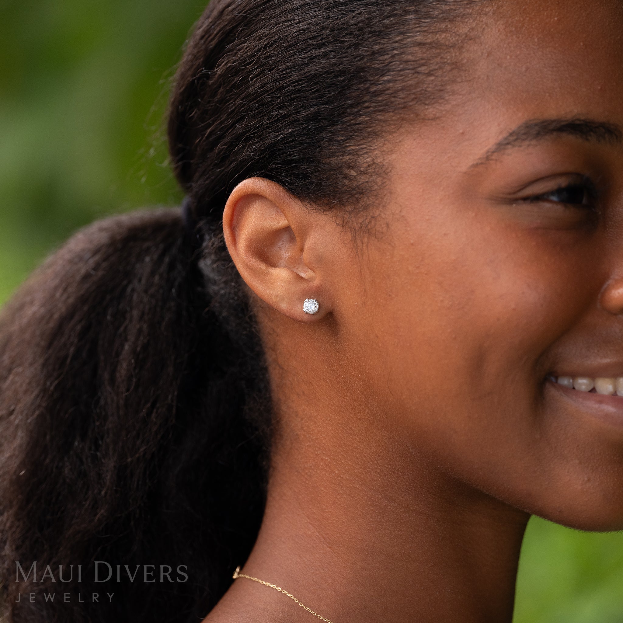 Close-up of a smiling woman wearing a Hidden Hawaiʻi Maile Lab Grown Diamond Earrings in 14k White Gold on her ear, against a blurred green background