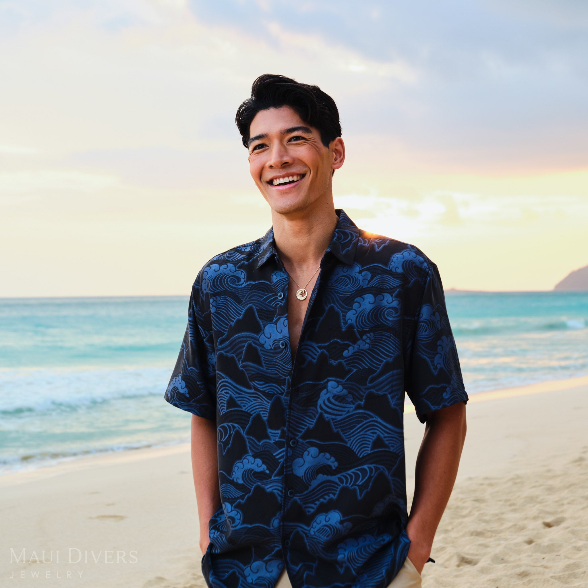 Man on a beach at sunrise wearing a dark blue patterned shirt with a gold diamond Nalue wave pendant