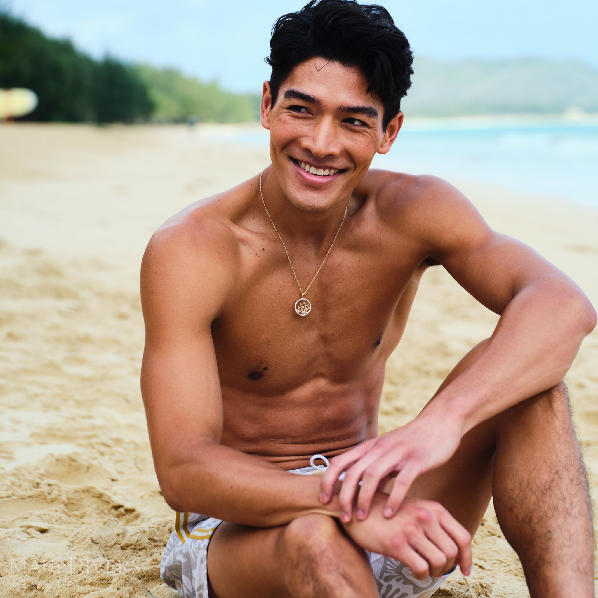 Man sitting on a beach wearing a gold coral branch pendant with diamonds on a rope chain