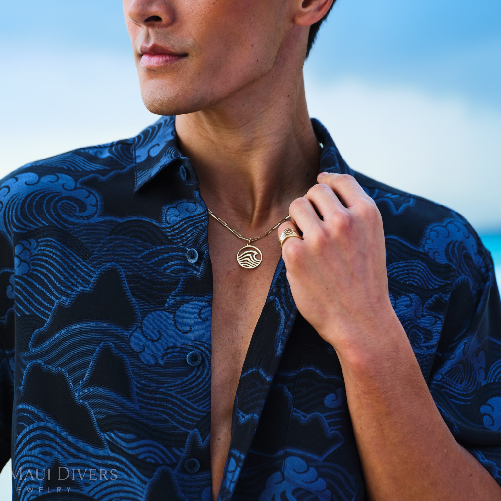 Man wearing a blue patterned shirt with a wave necklace and ring, against a blurred beach background.
