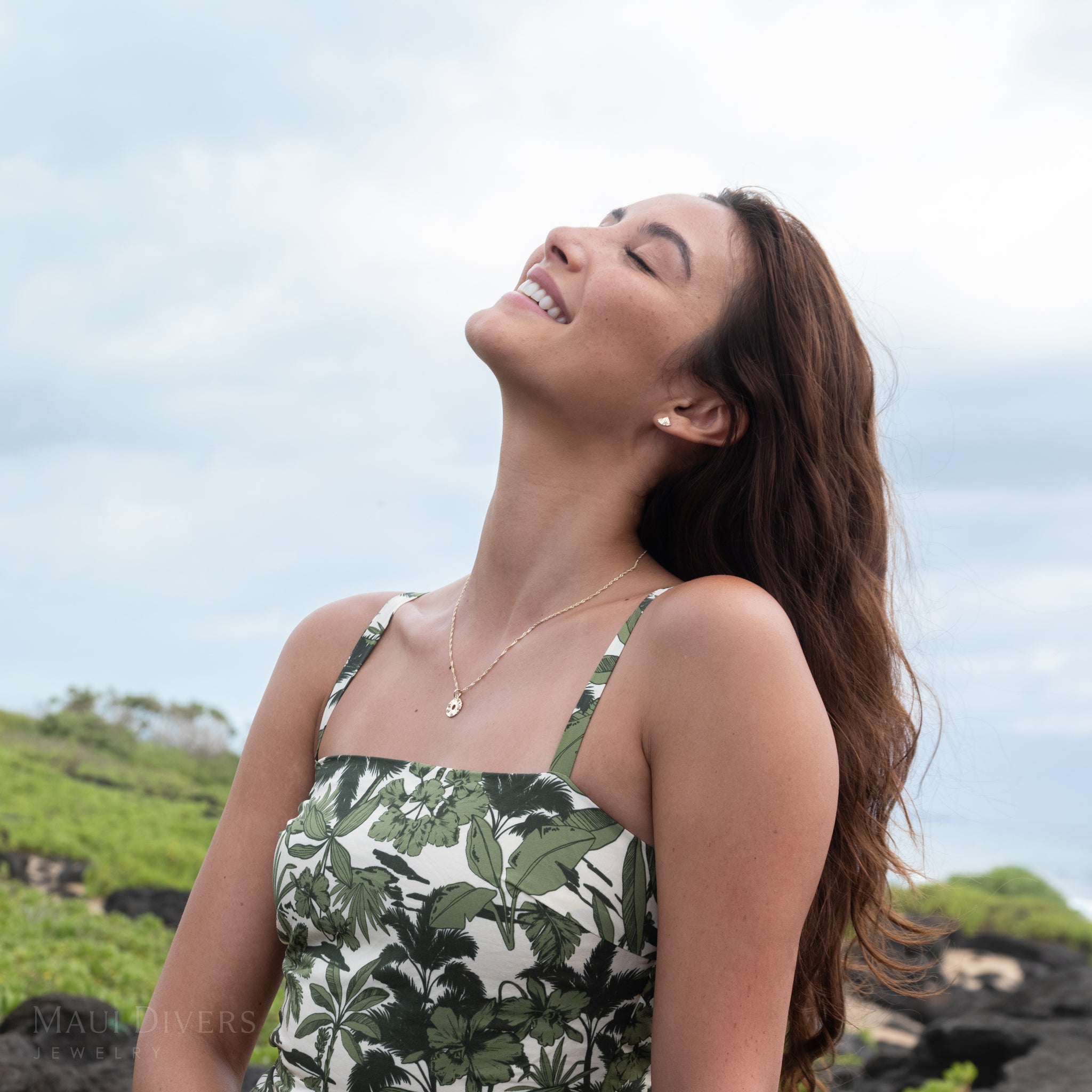 Woman wearing a round, gold and orange sapphire pineapple pendant on a singapore chain, orange sapphire pineapple slice earrings, and a patterned blue and green dress with a scenic green and sky background