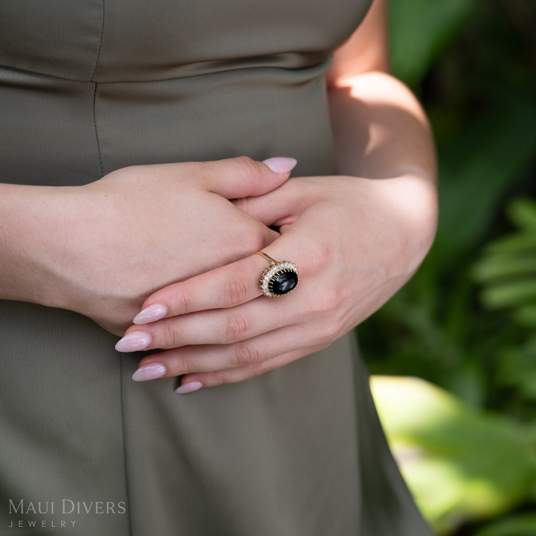 A close-up lifestyle shot of a model wearing a yellow gold ring with a large oval Hawaiian black coral cabochon and a halo of diamonds, set against an olive green dress and a blurred green background. The 'Maui Divers Jewelry' logo is visible in the bottom-left corner.