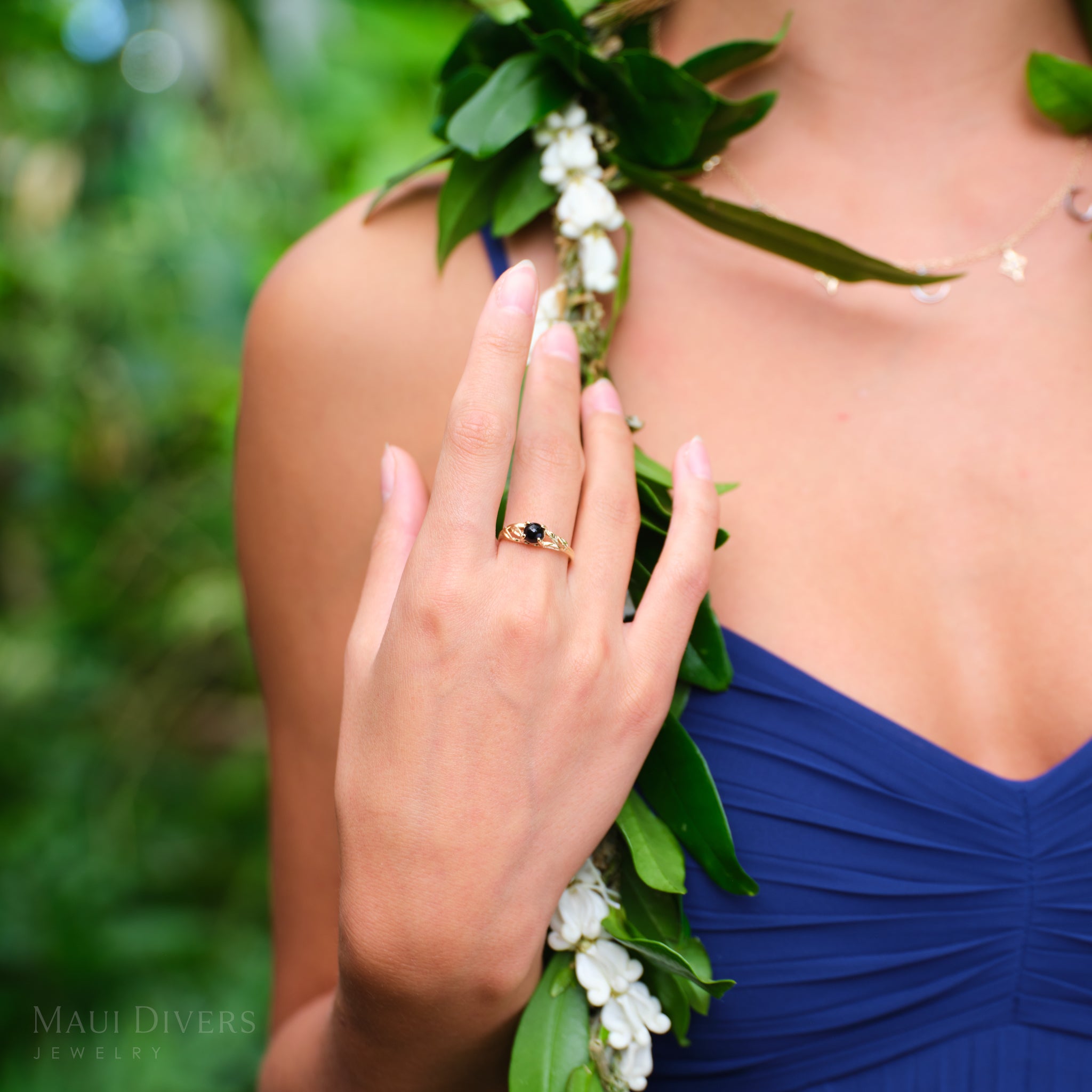 Hand wearing a Maile Leaf Black Coral Ring in 14k Yellow Gold over a maile lei with pikake flowers