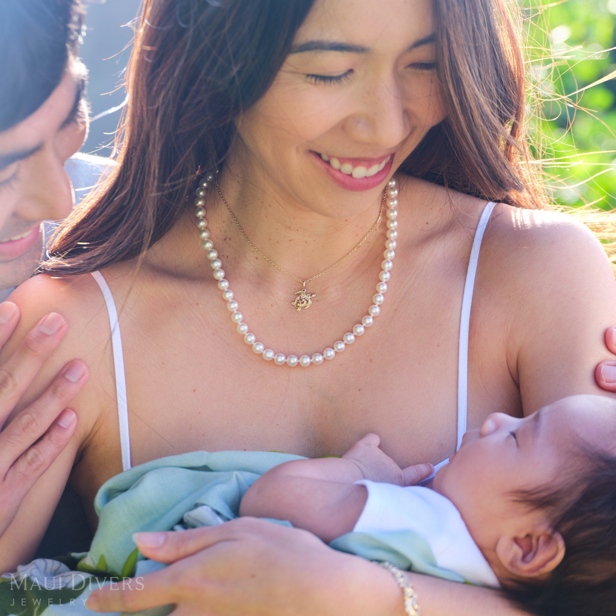 Smiling woman wearing a Living Heirloom Honu pendant in 14k yellow gold and a white pearl strand necklace, looking at a baby with a man smiling over her shoulder