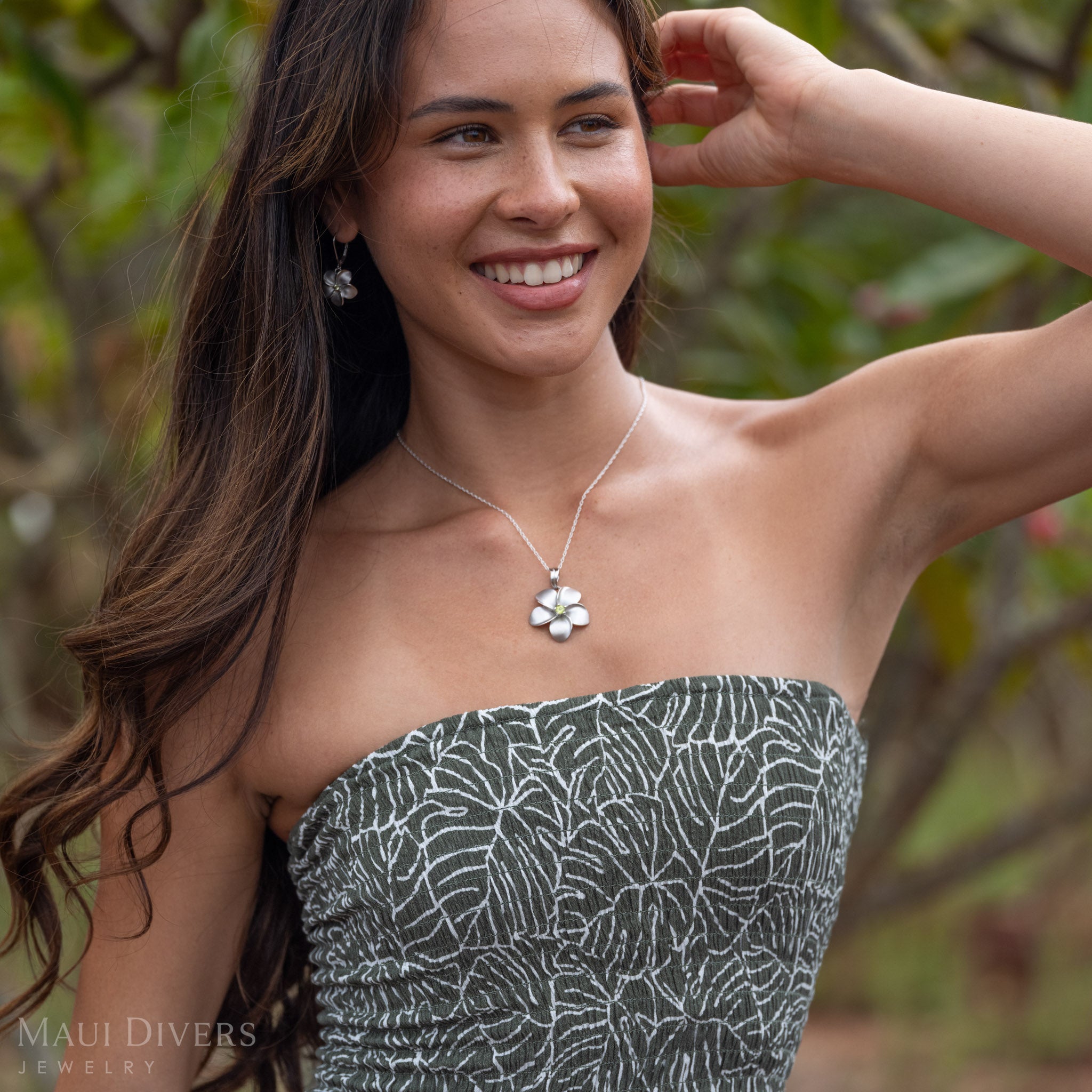 Woman smiling wearing Plumeria Peridot Necklace in Sterling Silver with matching earrings against a green background of plumeria trees.