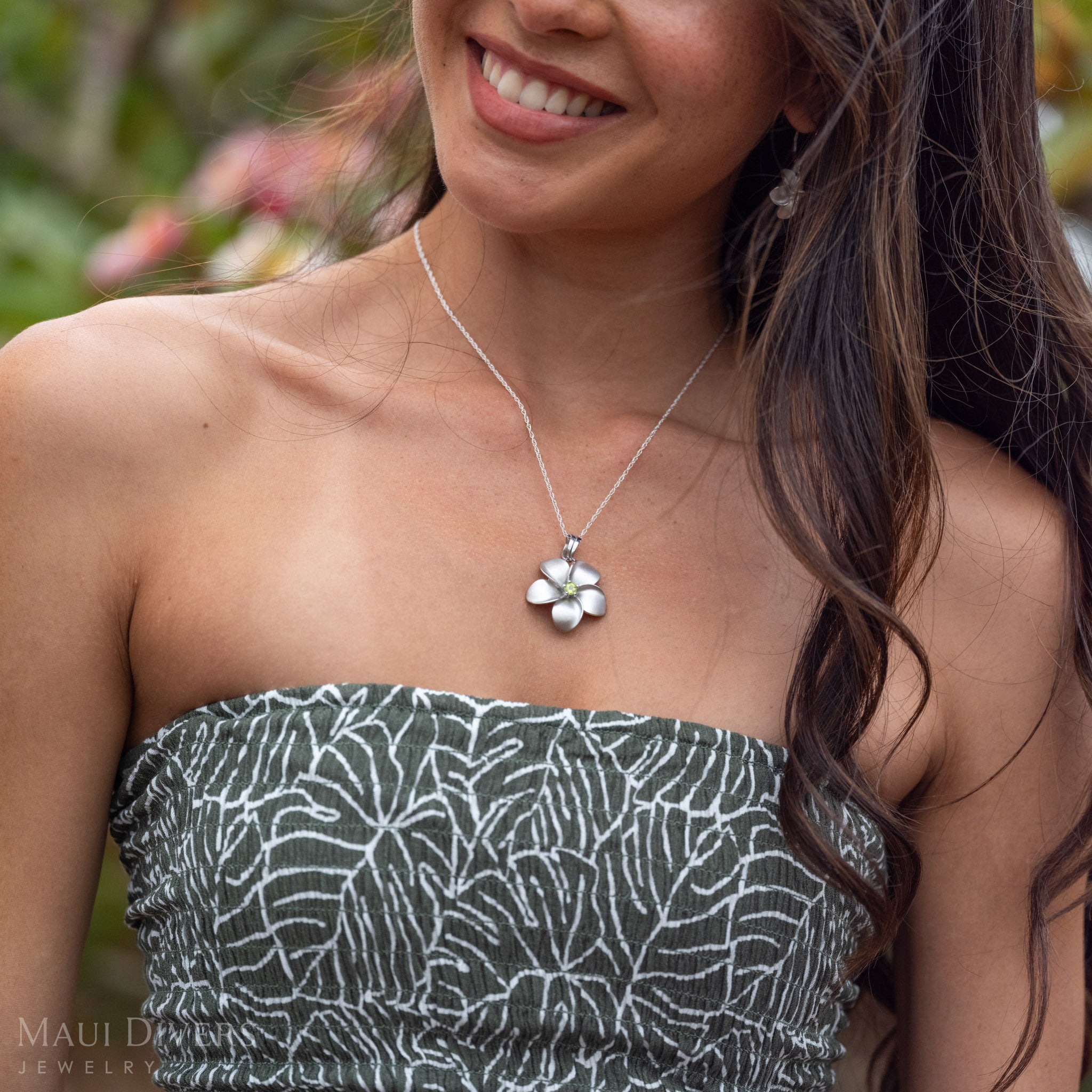 Woman smiling wearing Plumeria Peridot Necklace in Sterling Silver with matching earrings against a green background of plumeria trees.