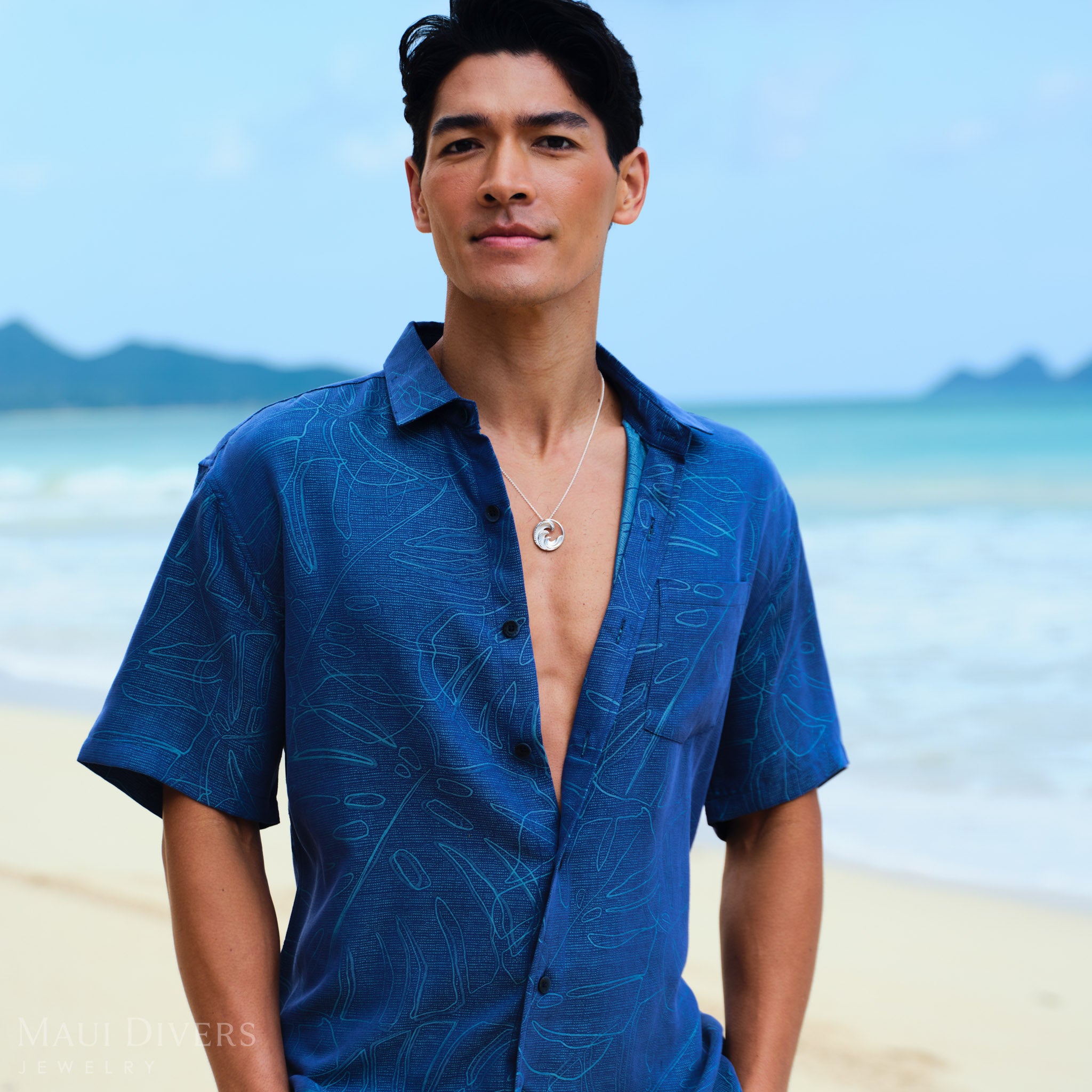 Man wearing a Nalu Pendant Necklace in Sterling Silver and blue patterned shirt on a beach