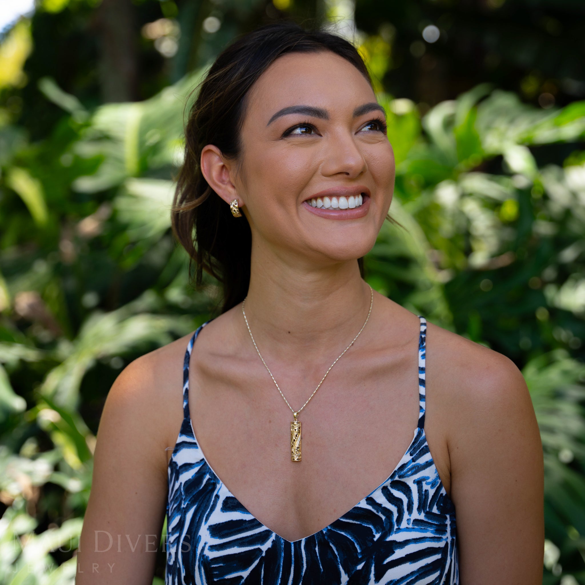 Woman wearing a Hawaiian Heirloom Plumeria Pendant in Gold standing in front of green foliage.