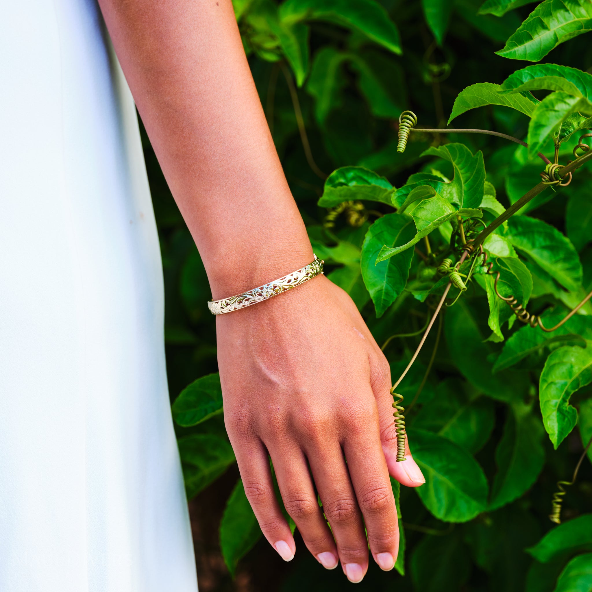 Hand wearing a Hawaiian Heirloom Plumeria Hinge Bracelet in Gold with green leaves in the background