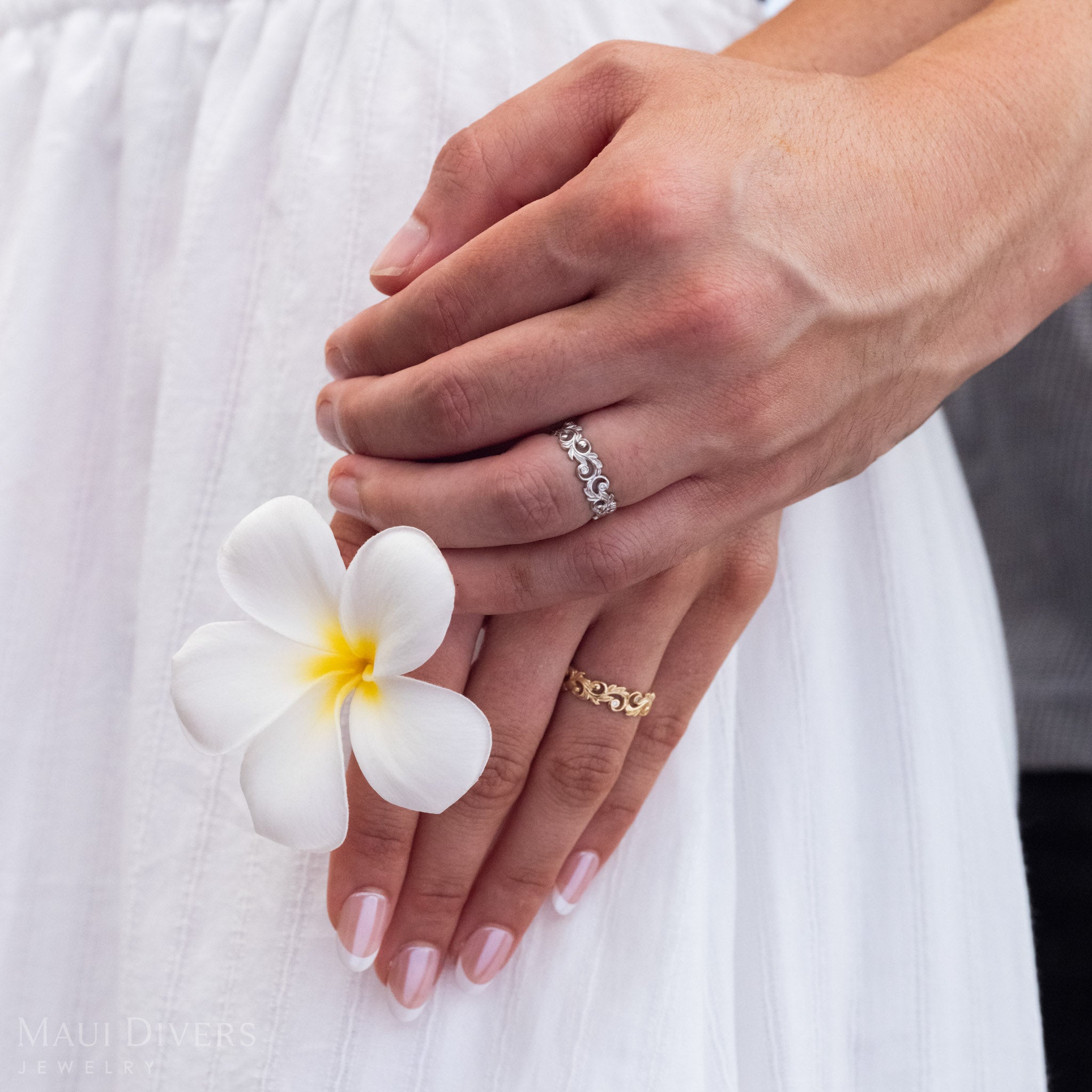 A hand holding another hand wearing Living Heirloom Ring in 14k White Gold and 14k Yellow Gold with Diamond accents, holding a plumeria flower.
