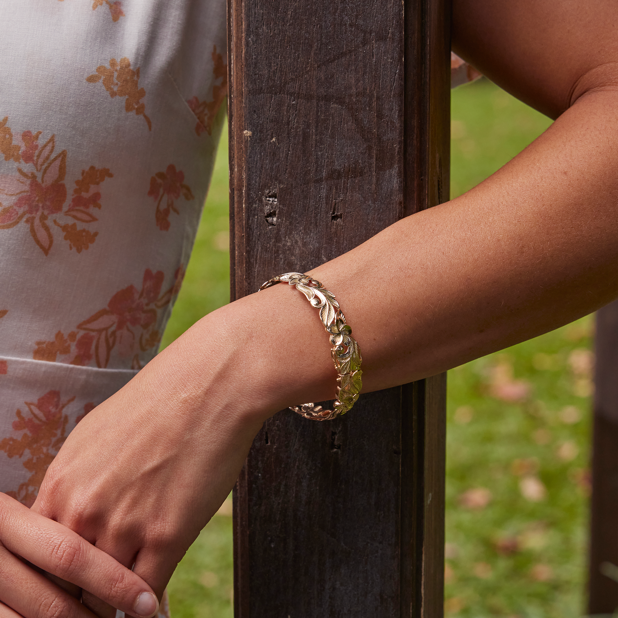 Hawaiian Heirloom Plumeria Hinge Bracelet in Gold on an arm wrapped around a brown post