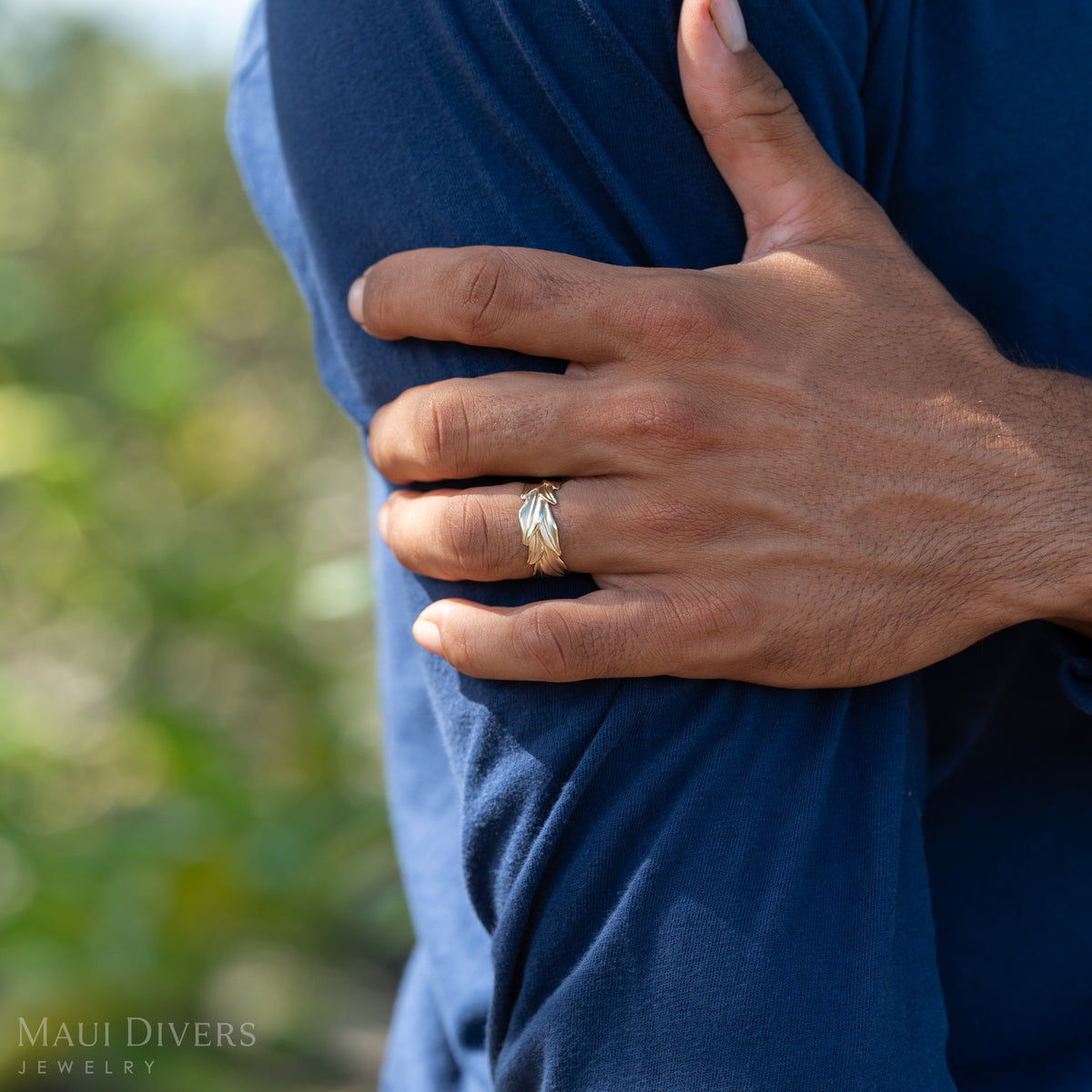 Close up shot of Dane Little's hand on his arm wearing a large gold Maile leaf ring against a blurry green background