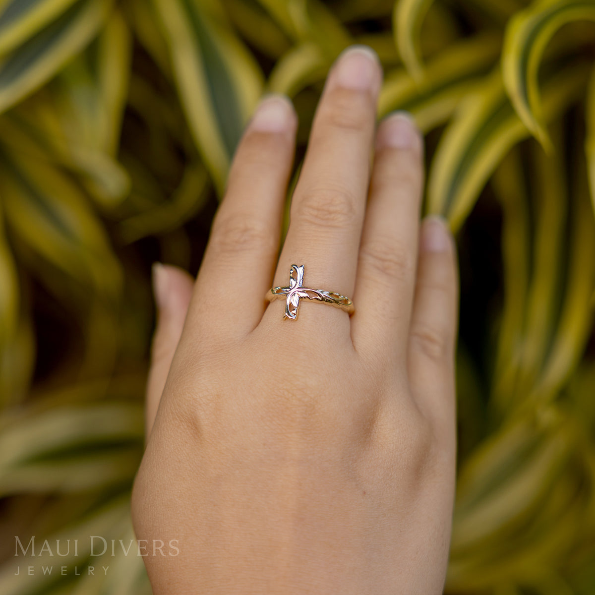 Close-up of a hand wearing a Hawaiian Heirloom Old English Scroll Cross Ring in 14k yellow gold, against a blurred leafy green background