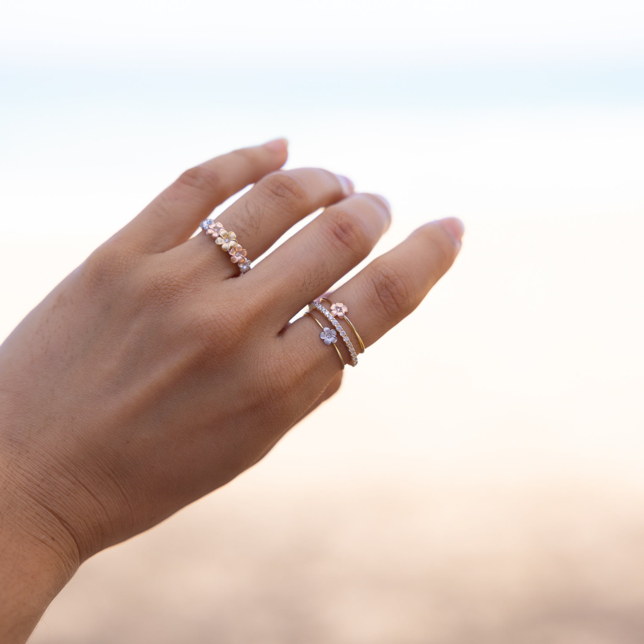 Hand wearing multiple Plumeria Rings in Gold with Diamonds against a blurred sand background