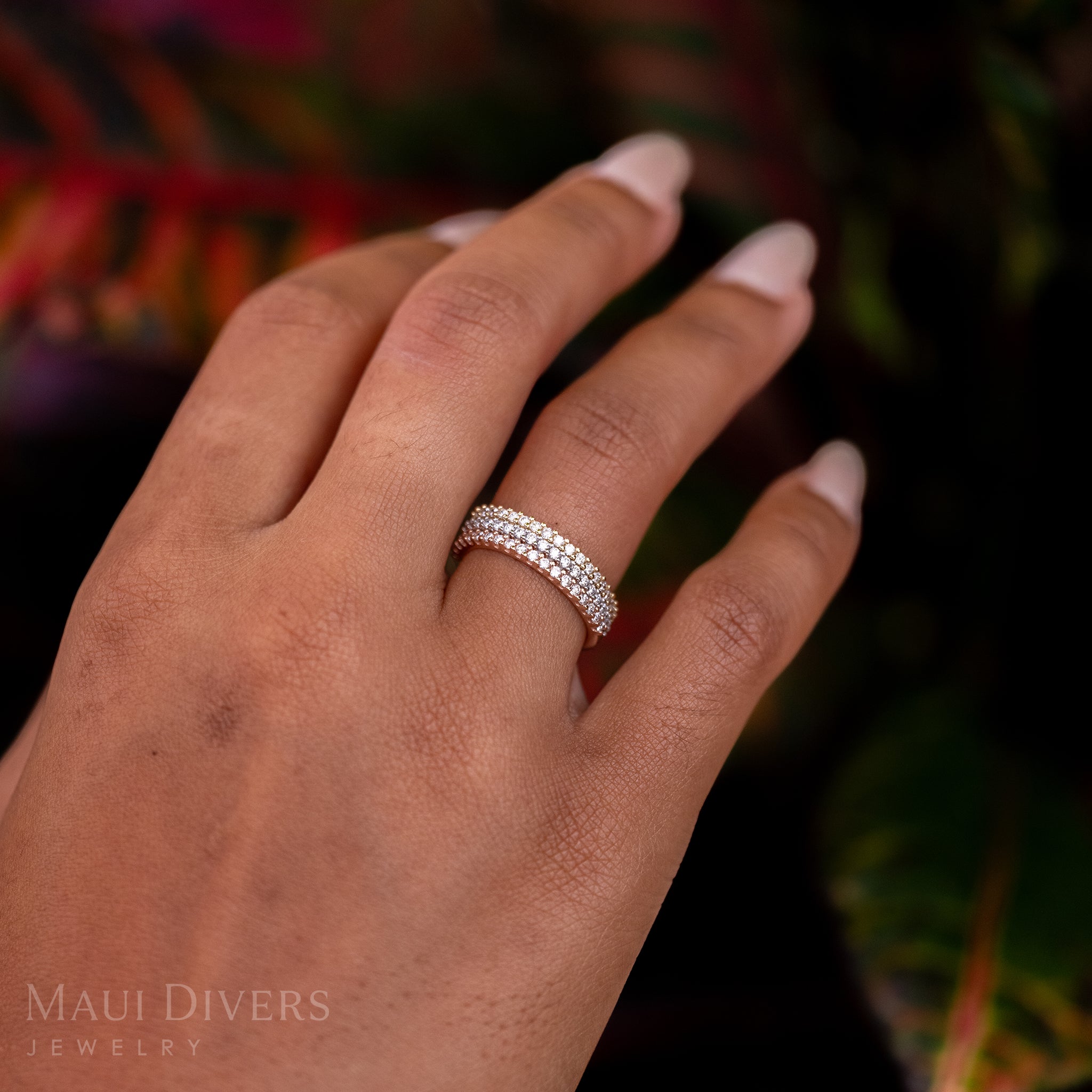 A close-up of a hand wearing a stack of Anniversary Rings in 14k gold, white, and rose gold with diamonds, against a blurred leafy green, red, and yellow background