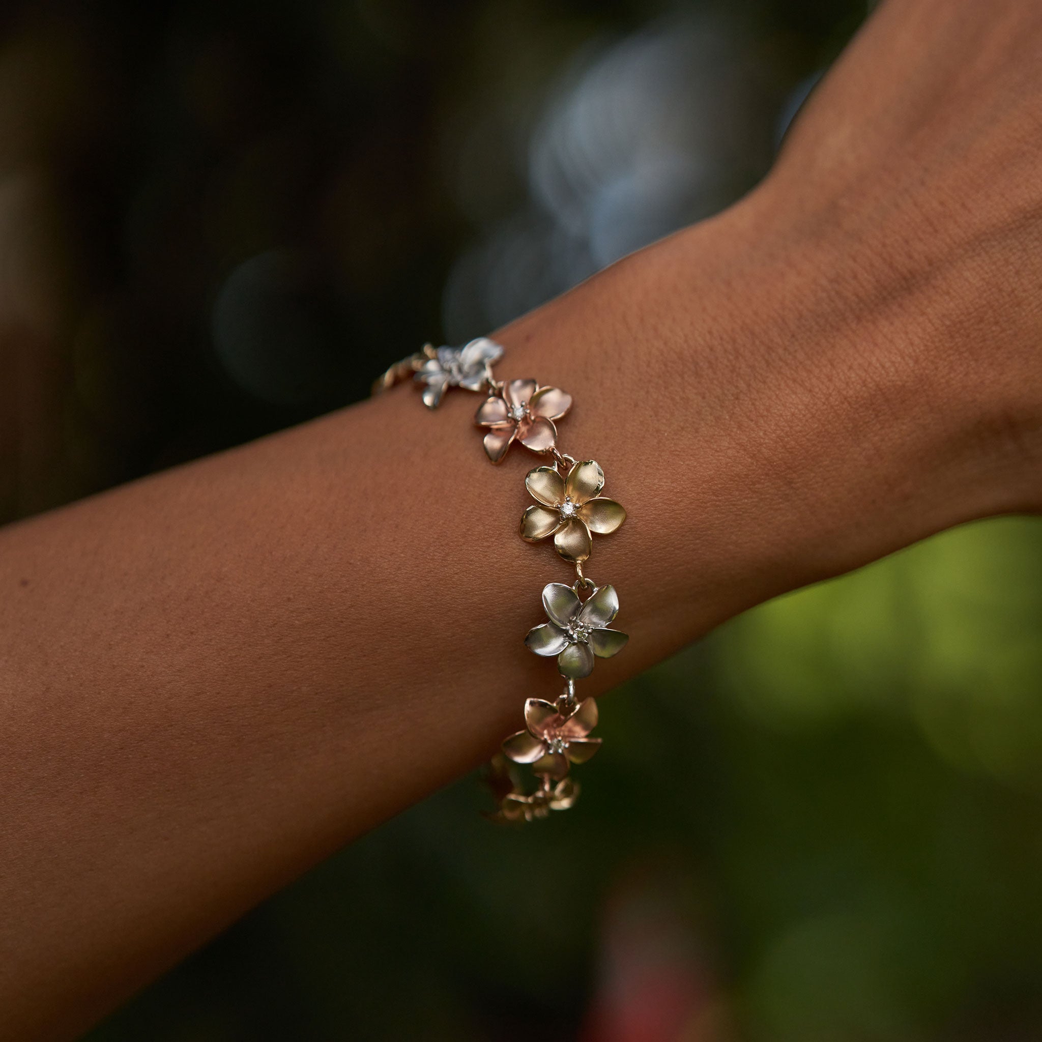 Close up of a 13mm Plumeria Link Bracelet in Tri Color Gold with Diamonds on a wrist against a blurred green background