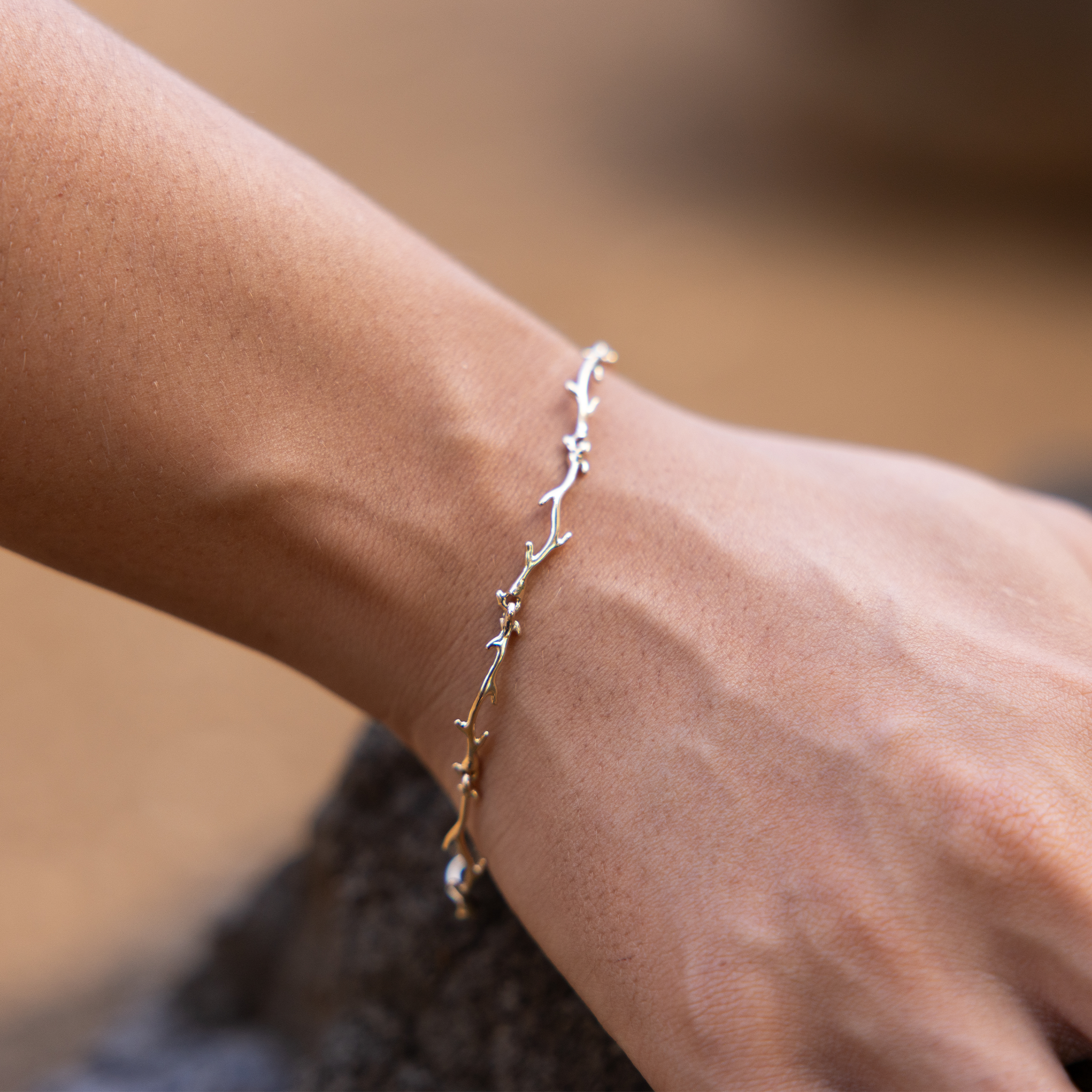 Close up of a Heritage Coral Branch Link Bracelet in 14k Yellow Gold with blurred rock and sandy background