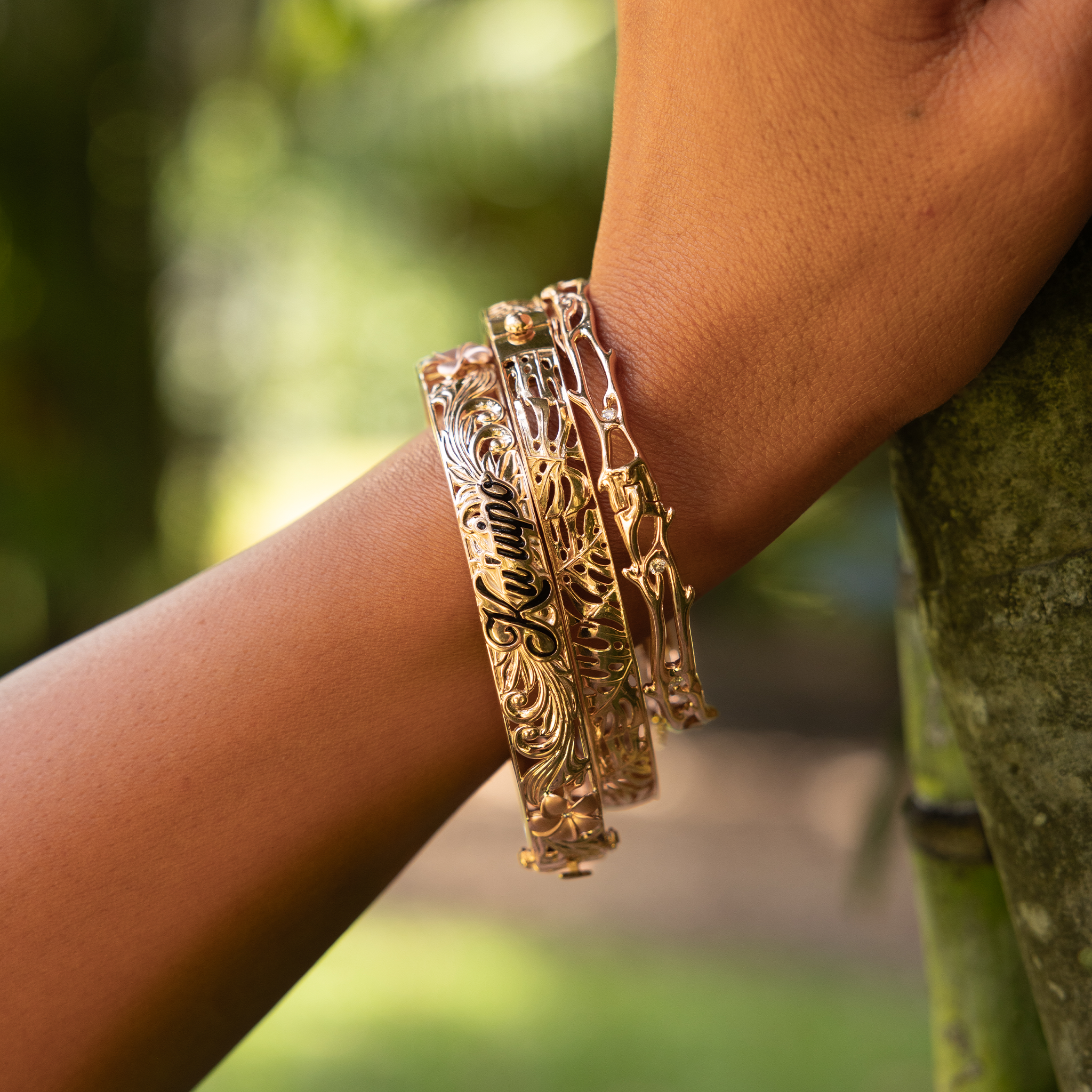 Close up of Monstera, Heritage and Hawaiian Heirloom Plumeria Hinge Bracelets in Gold on wrist outdoors