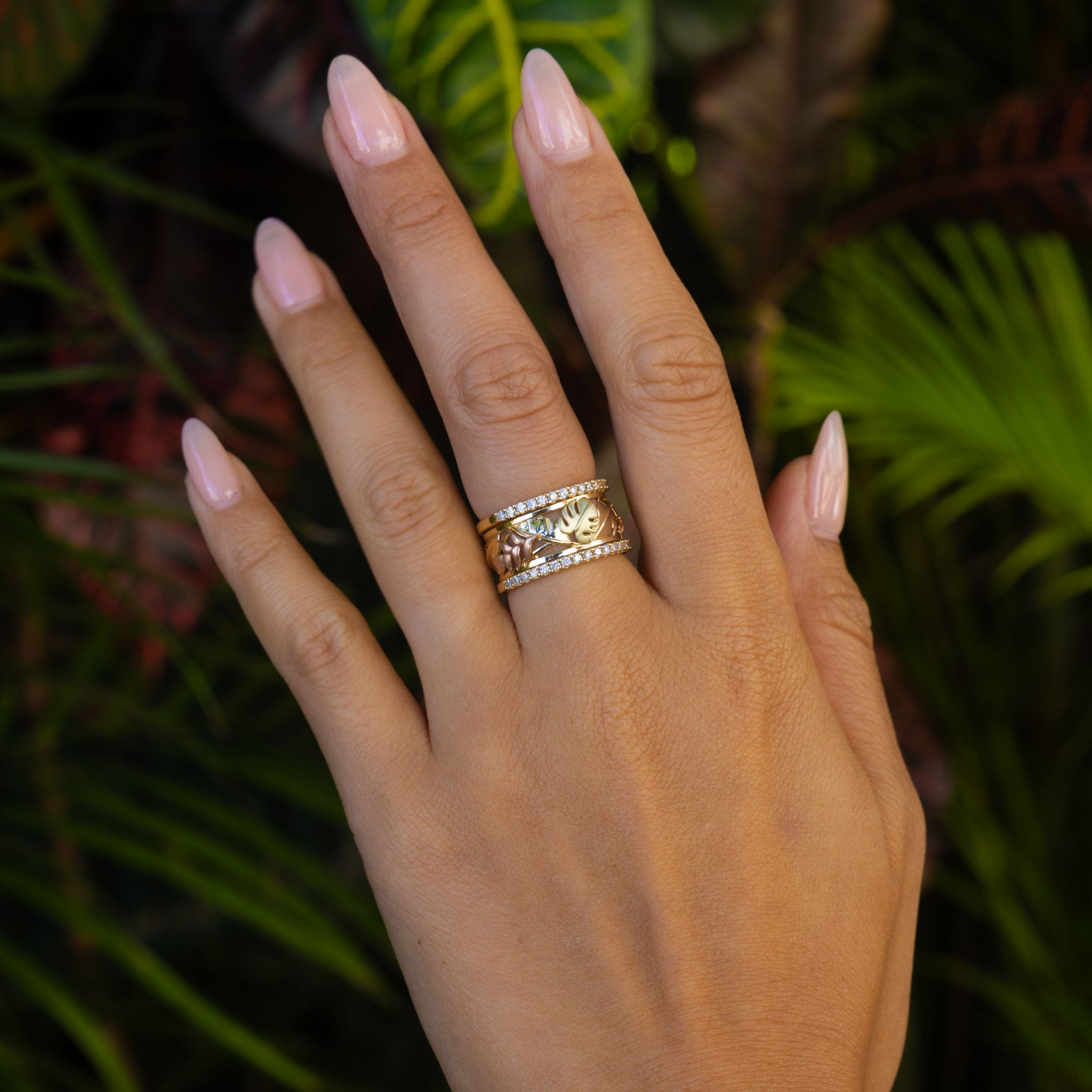 Hawaiian Gardens Monstera & Heliconia Ring in Tri Color Gold with 2 Diamond Bands on hand over blurred leaves