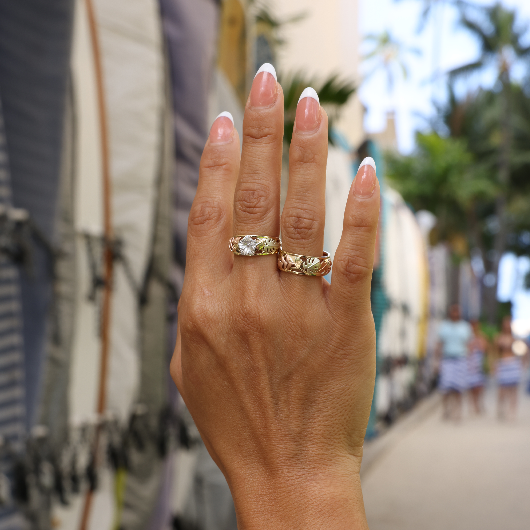 Two Hawaiian Gardens Monstera & Heliconia Rings in 4 Color Gold held up with surfboards blurred in the background