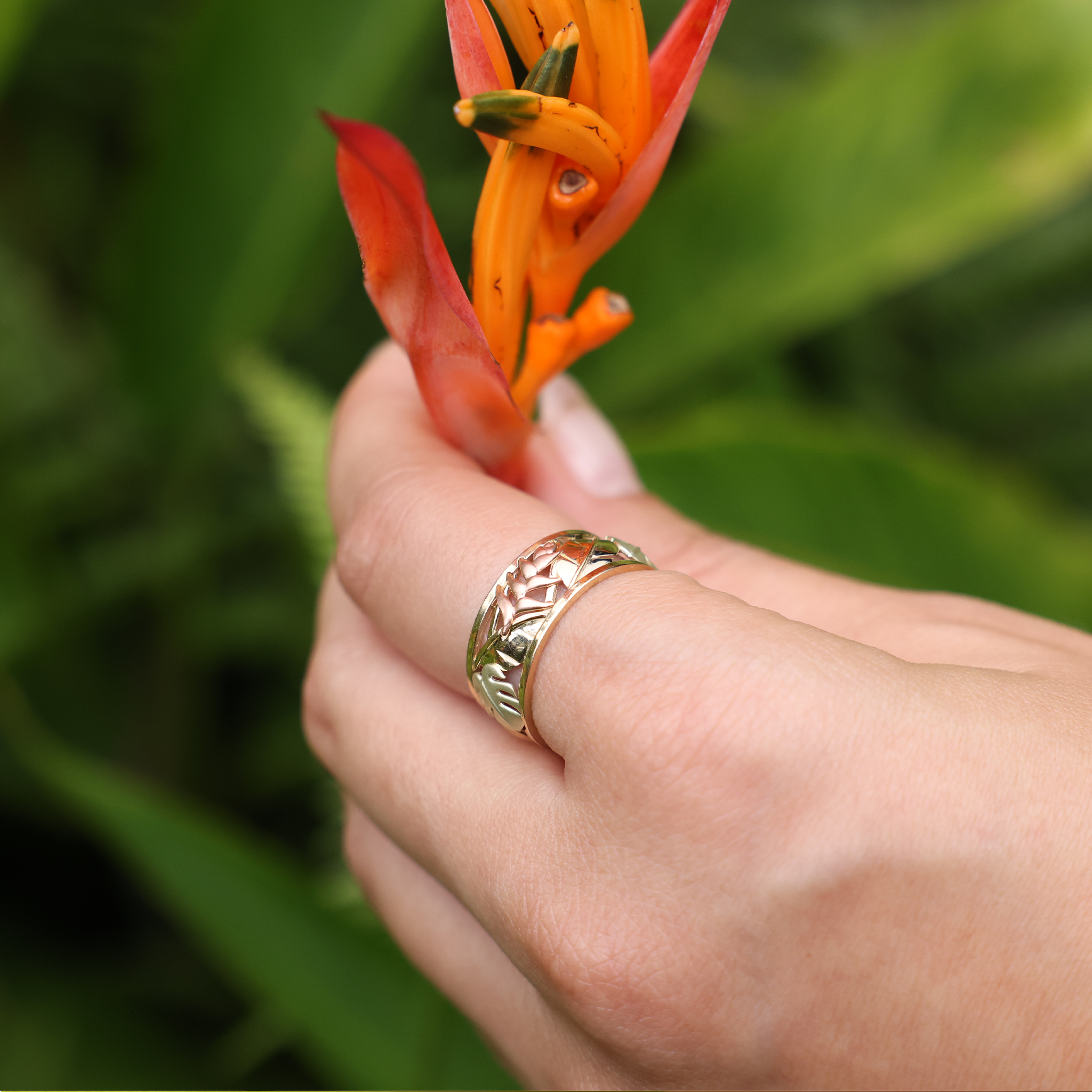 A womanʻs hand with a Hawaiian Gardens Monstera & Heliconia Ring in Tri Color Gold holding a flower