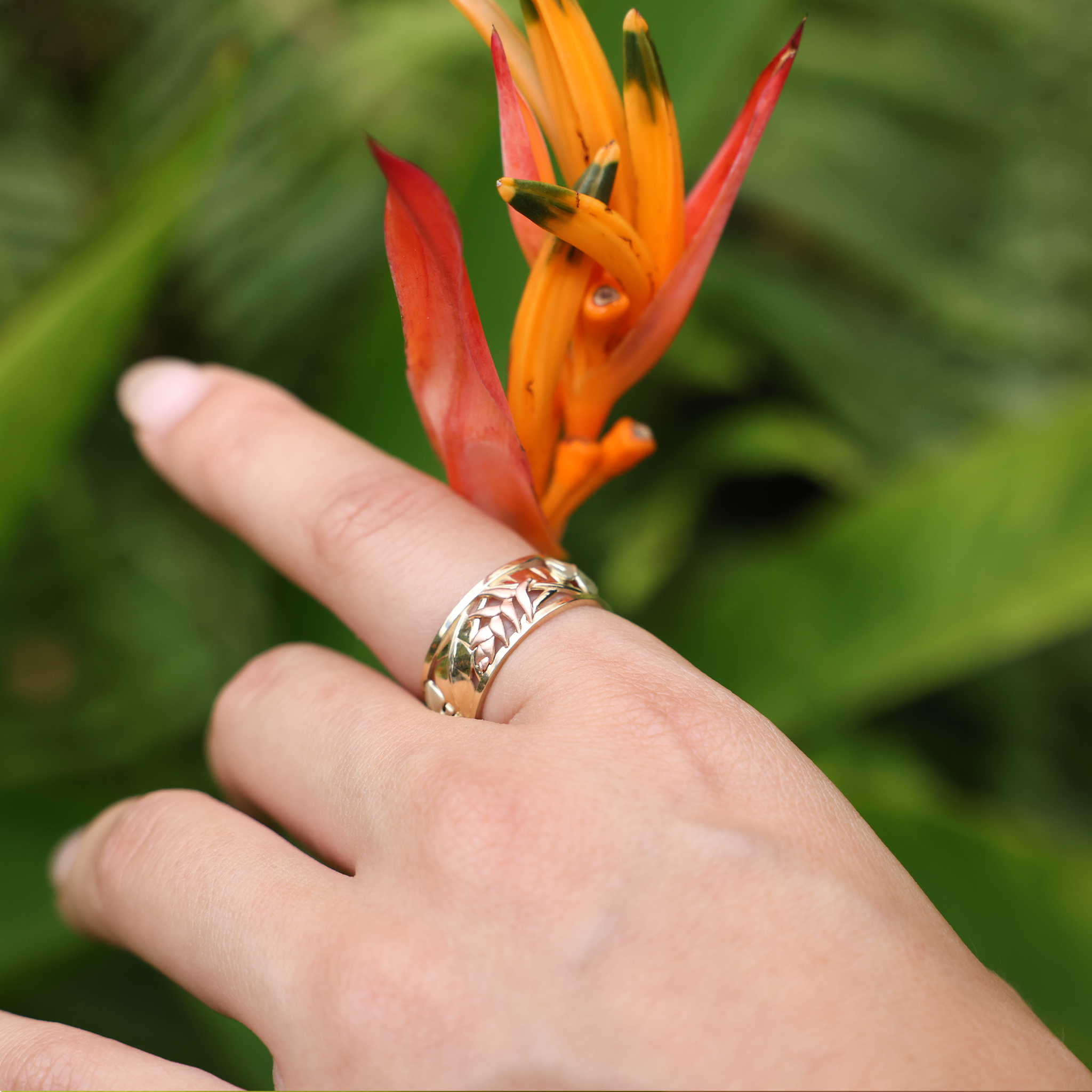 A woman's hand with a Hawaiian Gardens Monstera & Heliconia Ring in Tri Color Gold holding a flower