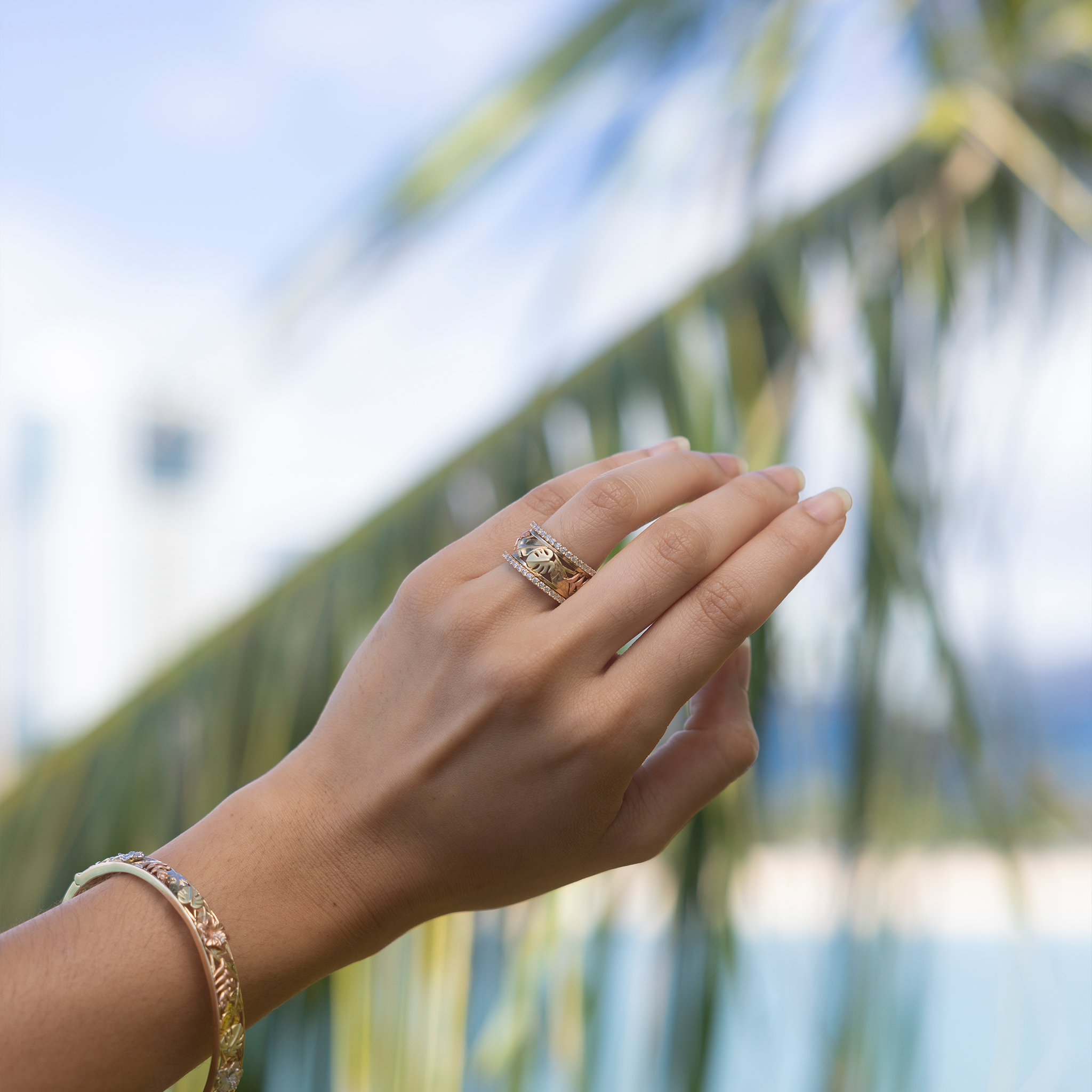 Hawaiian Gardens Monstera & Heliconia Ring in Tri Color Gold with 2 diamonds bands on hand held up against palm trees