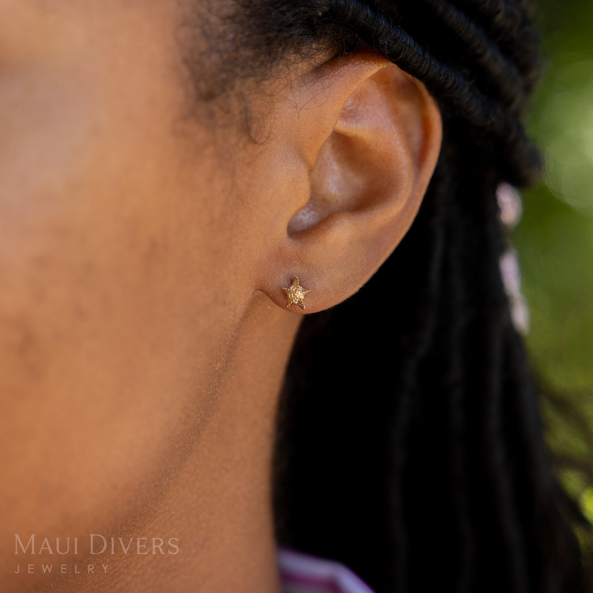 A side view close-up of a woman wearing a Starfish Earring in 14k yellow gold, against a blurred green background