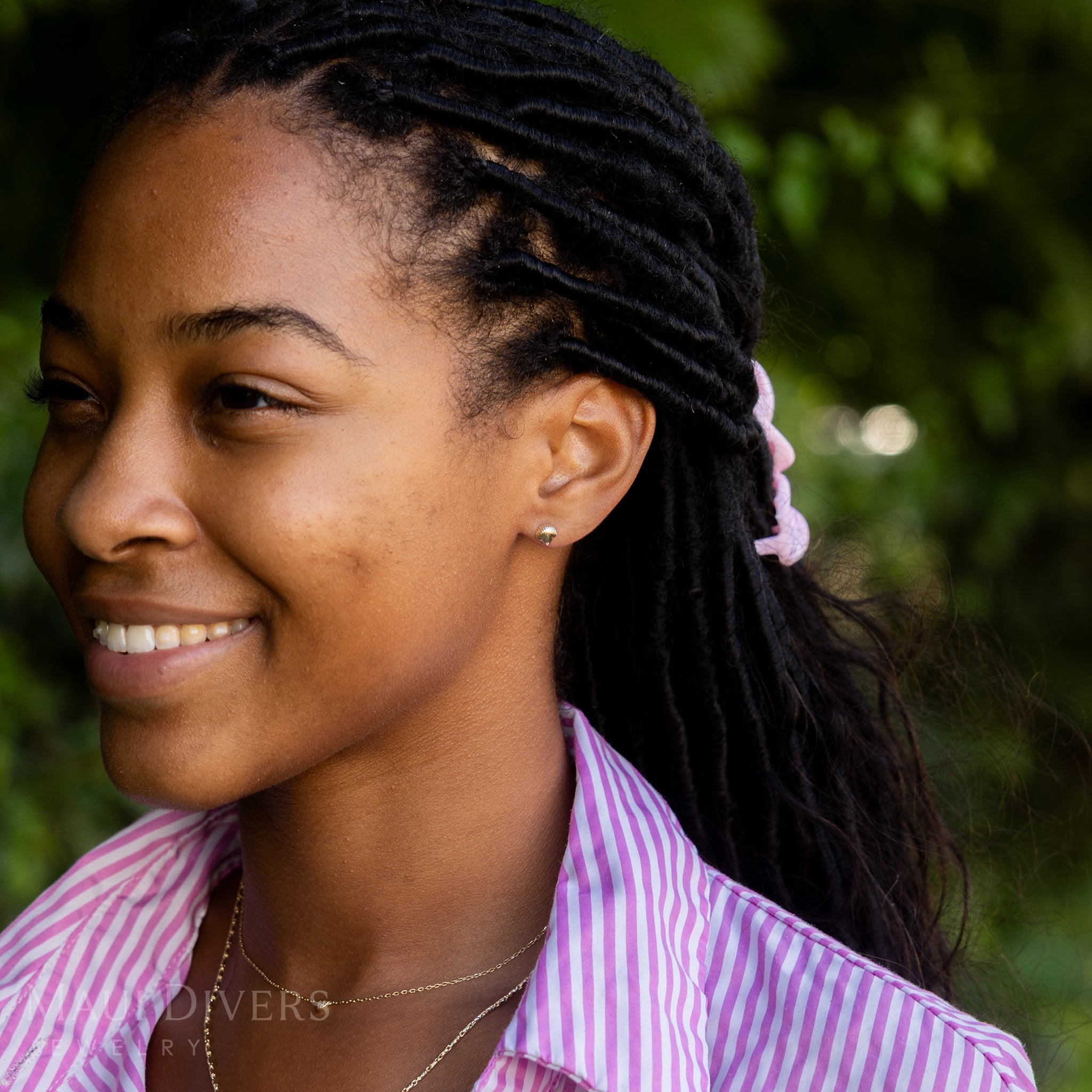 A side view of a smiling woman in a pink and white striped top wearing a Scallop Shell Earring in 14k yellow gold, paired with a tiny diamond heart necklace, against a blurred leafy green background