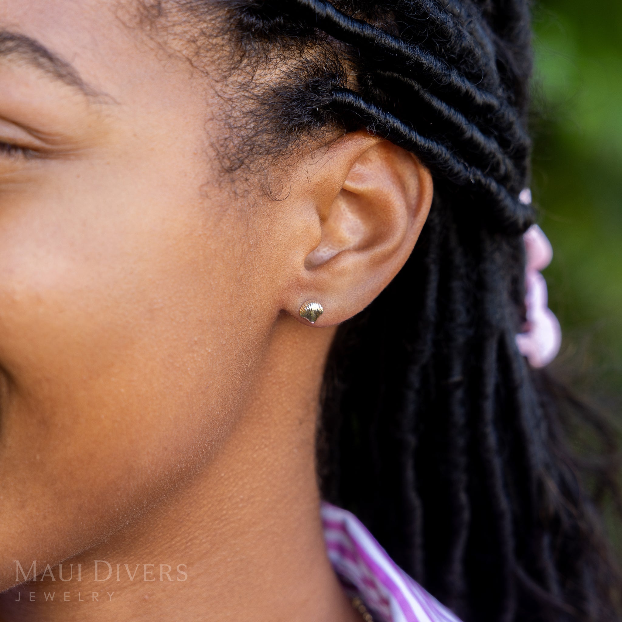 A side view close-up of a smiling woman in a pink and white striped top wearing a Scallop Shell Earring in 14k yellow gold, against a blurred leafy green background