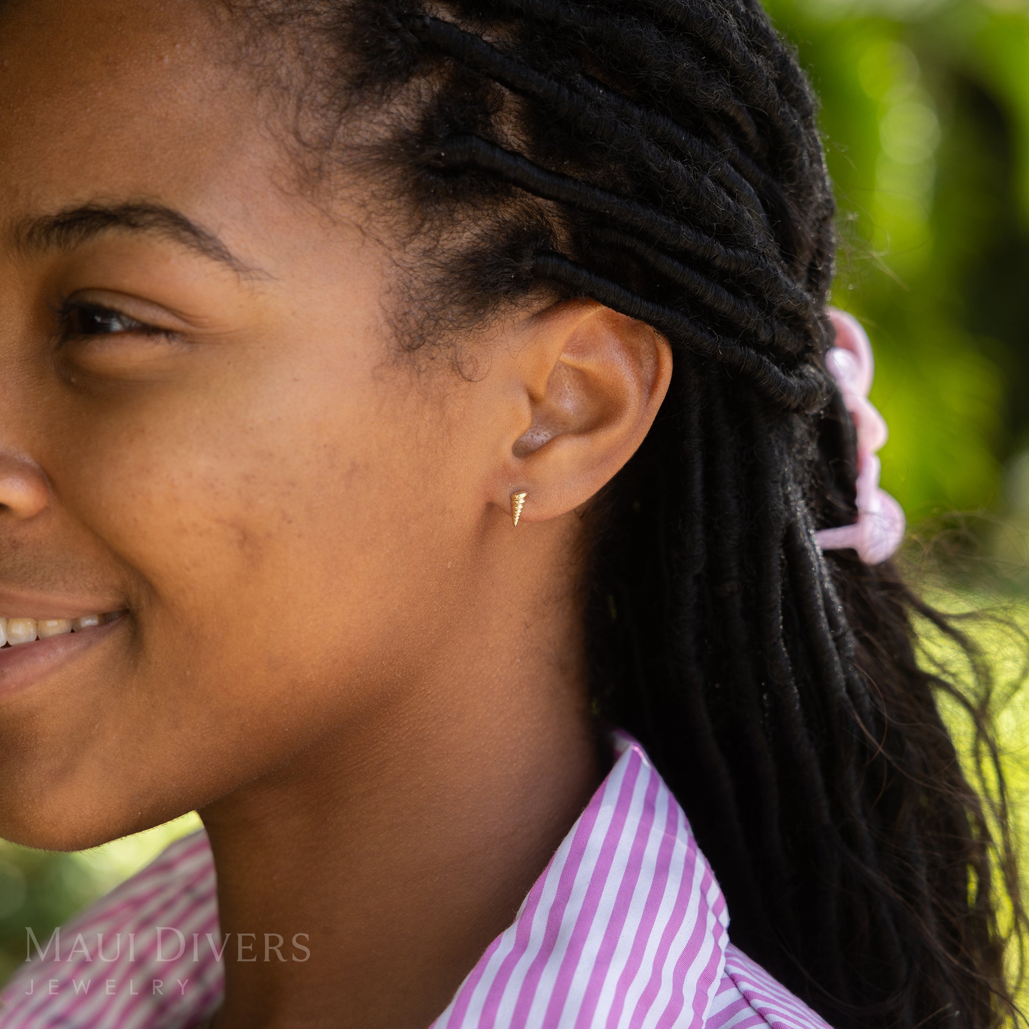 A side view of a smiling woman in a pink and white stripe top wearing a Conch Shell Earring in 14k yellow gold, against a blurred leafy green background