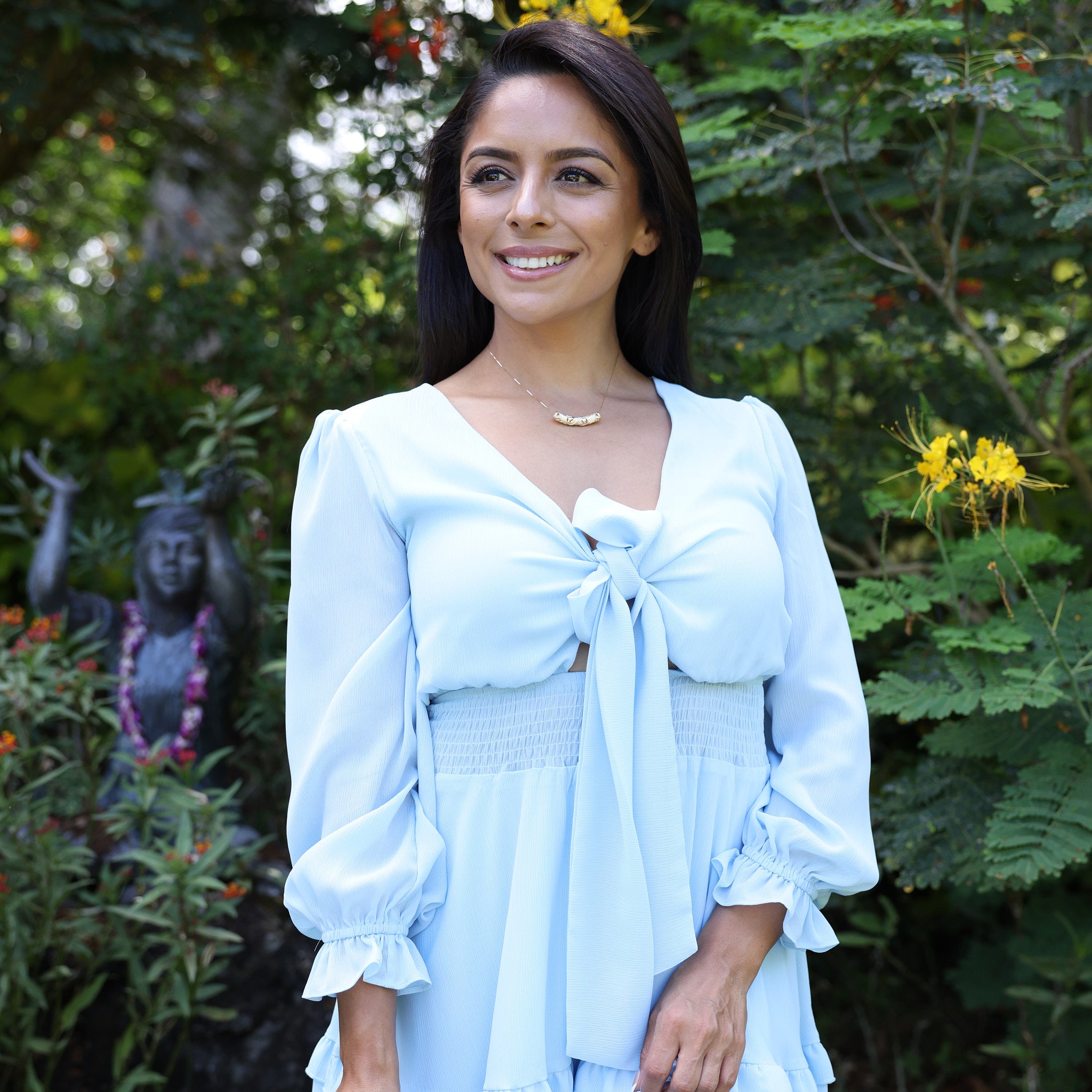 woman in blue dress wearing Hawaiian Heirloom Plumeria Barrel Pendant in Gold - 37mm in tropical botanical garden