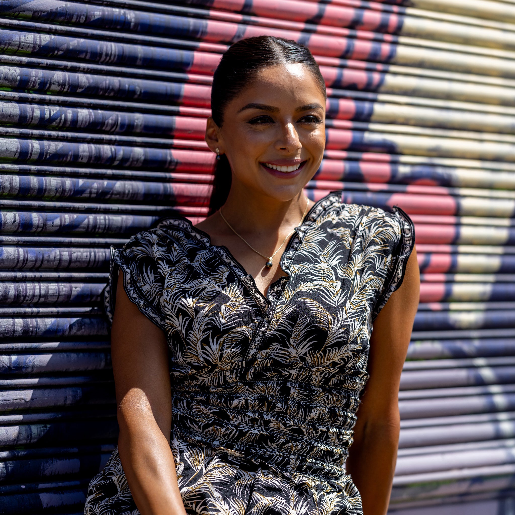 Woman in a black patterned dress wearing the Living Heirloom Tahitian black pearl pendant in 14k yellow gold with diamond accents on a 14k yellow gold chain with a graffiti backdrop