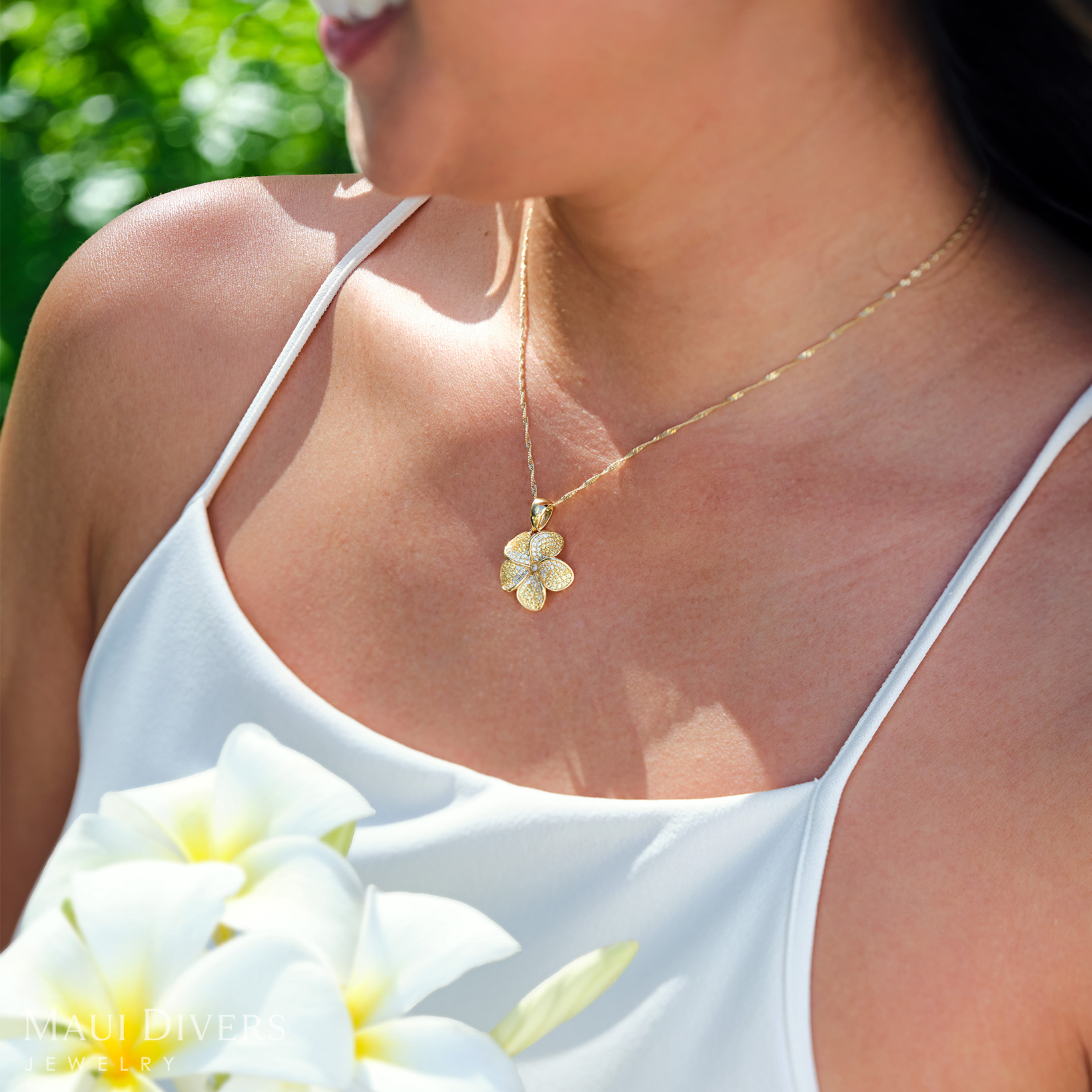 Woman wearing a Plumeria Yellow Sapphire Pendant in Gold with Diamonds, holding white Plumeria flowers outdoors.